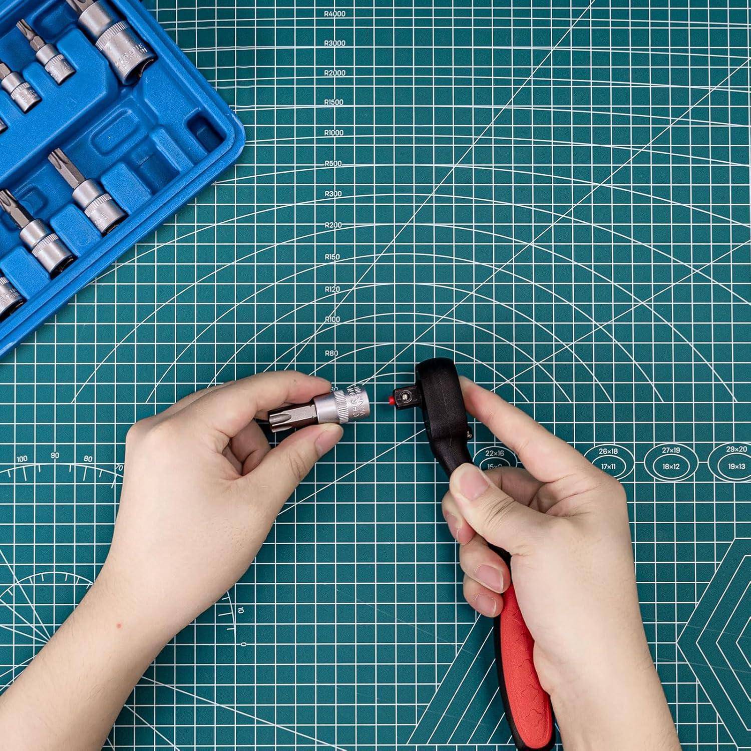 Hände verwenden eine Winkelgetriebevorsatzeinheit an einem Elektroschleifergerät, um einen Aufsatz auf einer grünen Schneidematte anzubringen, mit einem blauen Steckschlüsselsatz in der Ecke.