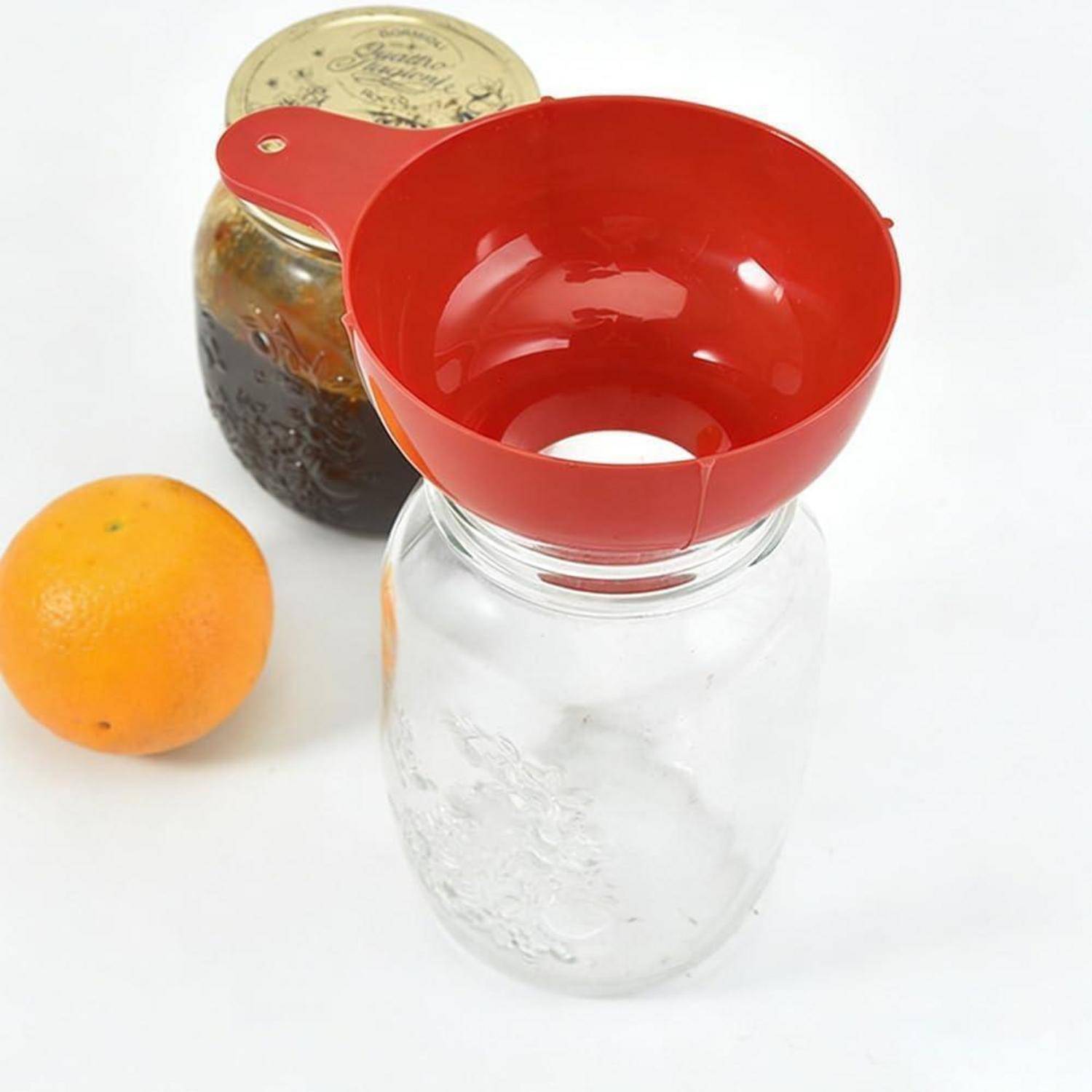 Roter Trichter auf einem Glaskrug mit einem Krug dunkler Flüssigkeit und einer Orange daneben auf weißem Hintergrund.