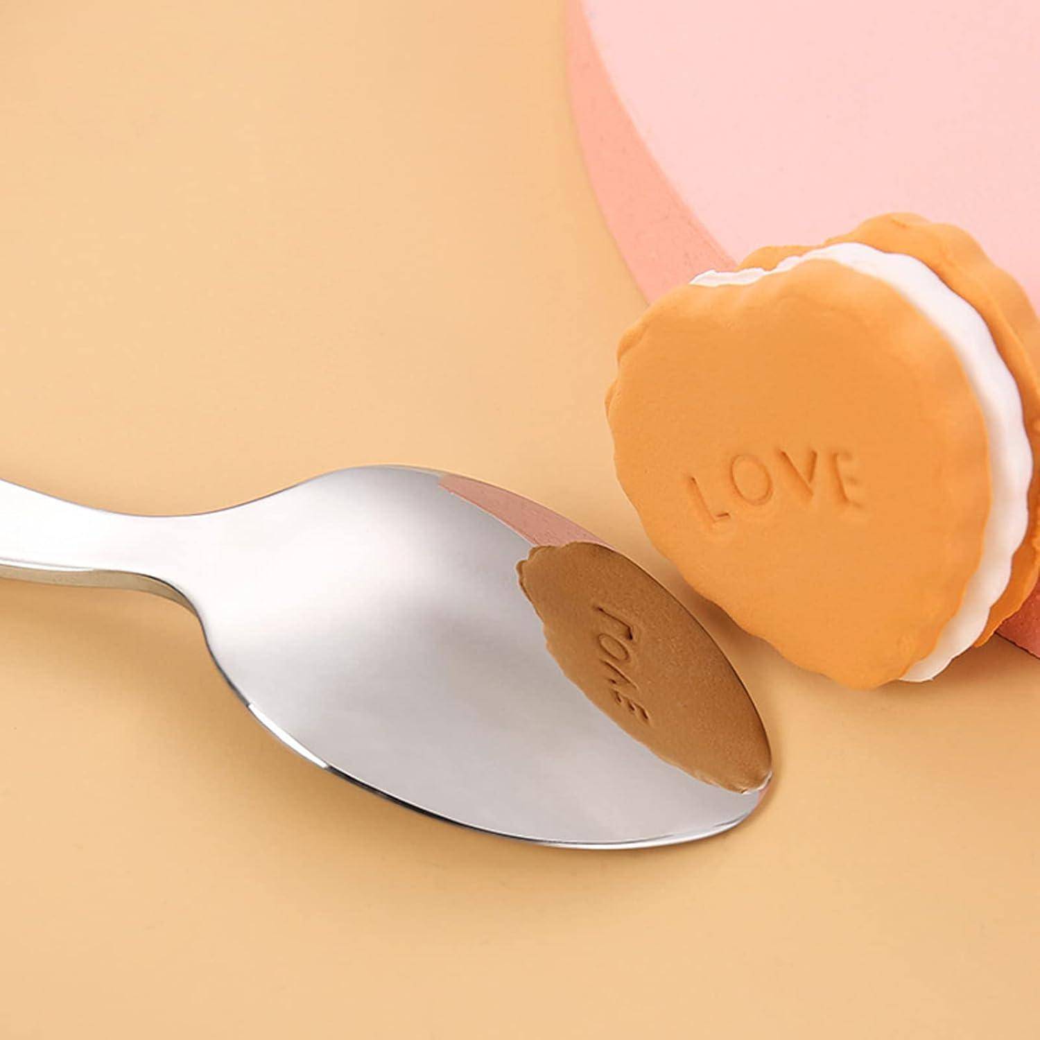 Ein Metalllöffel mit einem herzförmigen Keks, auf dem "LOVE"" geprägt ist, ruht neben einem rosa Behälter auf einem pfirsichfarbenen Hintergrund.