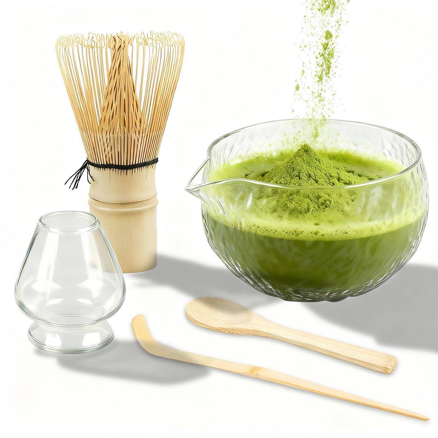 Matcha-Tee-Set mit Bambusbesen, Löffel, Glassschüssel mit grünem Pulver, kleiner Glastasse und Holzutensilien auf weißem Hintergrund.