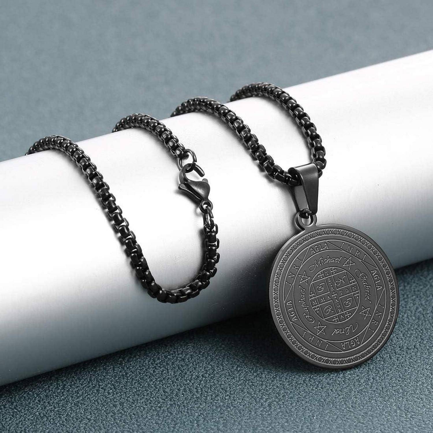 Schwarze Kettenkette mit einem runden gravierten Anhänger, ausgestellt auf einem weißen Zylinder vor einem grauen Hintergrund.