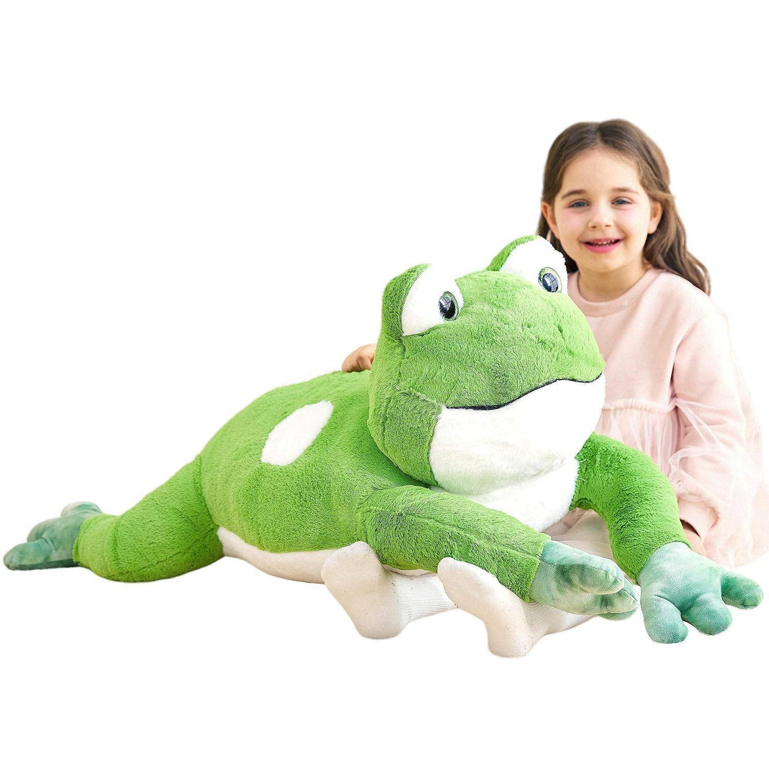 Junges Mädchen, das lächelt und ein großes grünes Frosch-Plüschtier auf weißem Hintergrund umarmt.