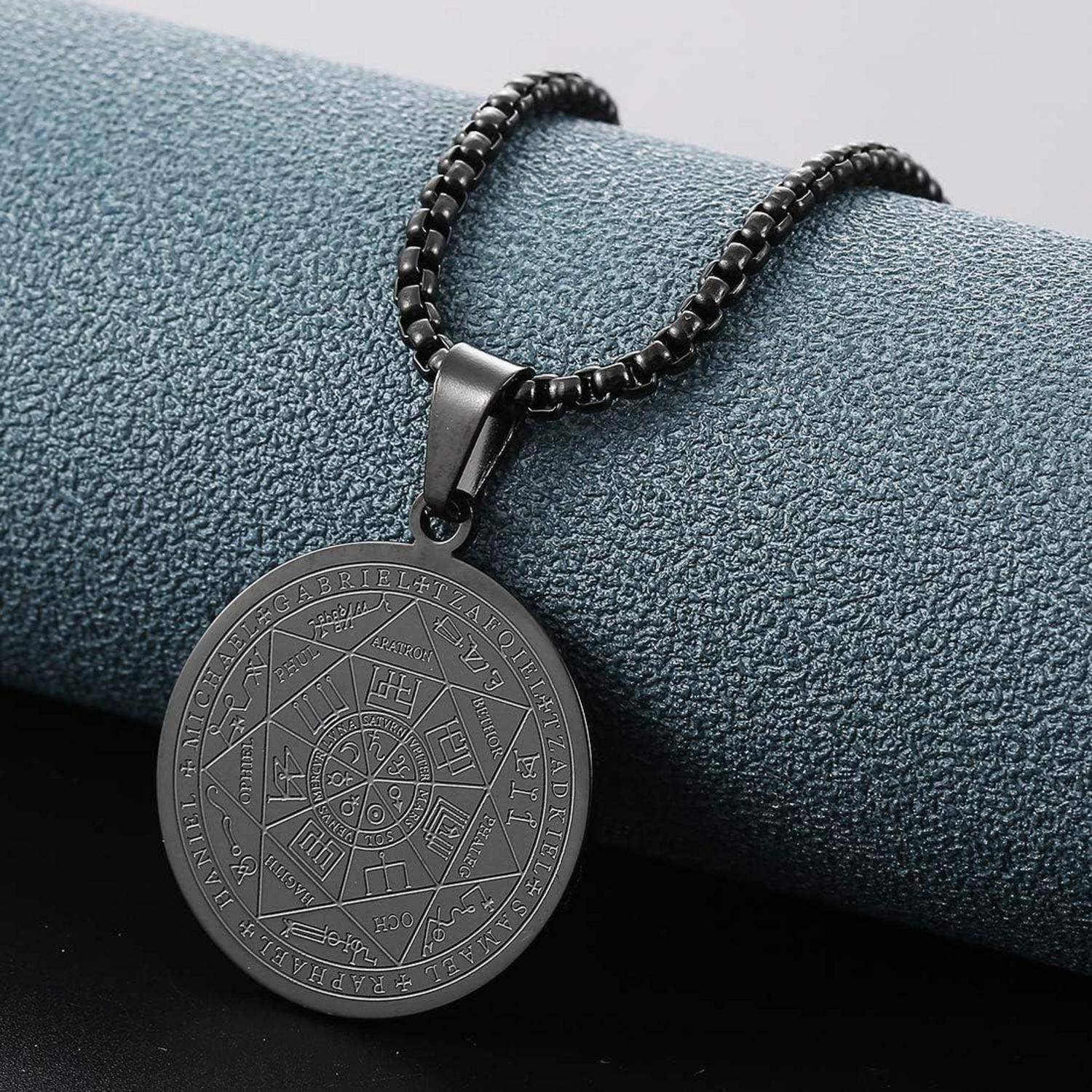 Schwarze kreisförmige Anhängerkette mit gravierten astrologischen Symbolen auf einer Perlenkette, ruhend auf einer strukturierten blauen Oberfläche.