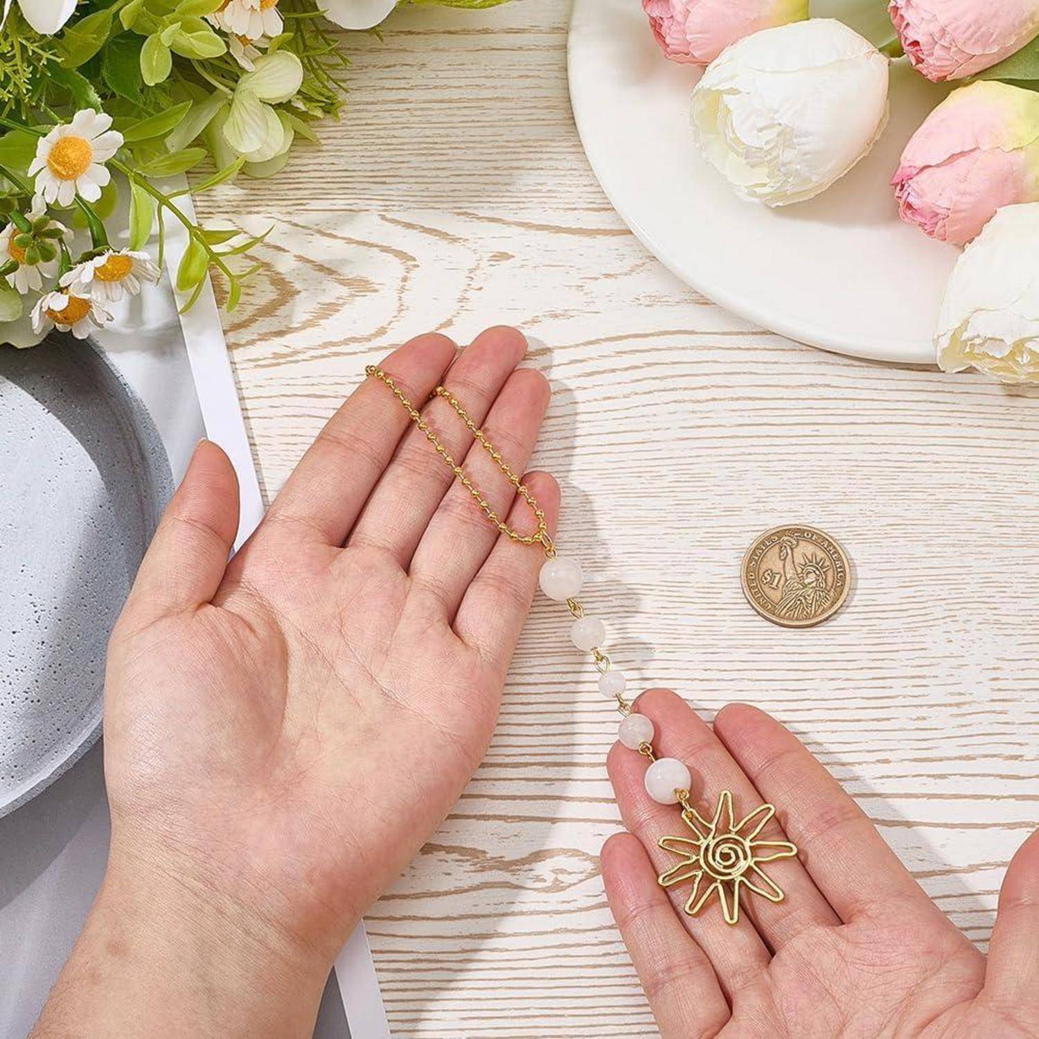 Hände halten eine goldene Perlenkette mit einem Blütenanhänger, ausgestellt auf einem hellen Holztisch mit Blumen und einer Münze als Größenvergleich.