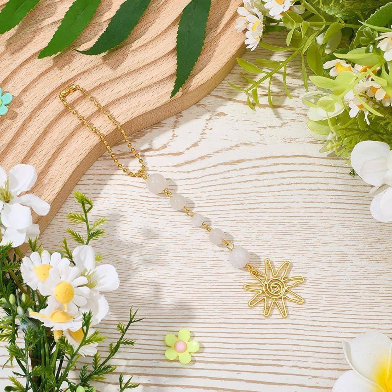 Goldene Perlenkette mit einem sonnenförmigen Anhänger auf einer hellen Holzoberfläche gelegt, umrahmt von weißen und gelben Blumen.