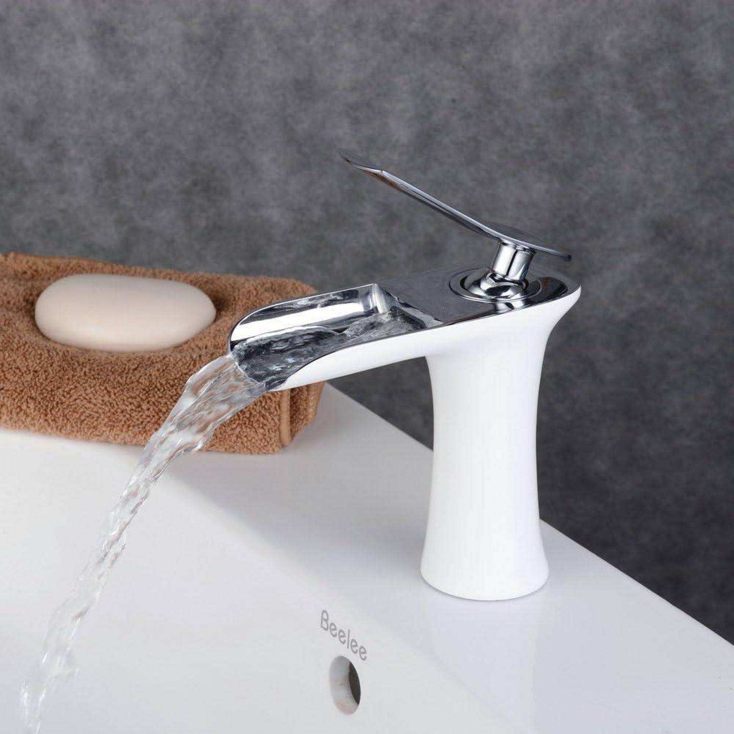 Moderner weißer Badezimmerwasserhahn gießt Wasser in ein weißes Becken neben einem braunen Handtuch mit Seife.