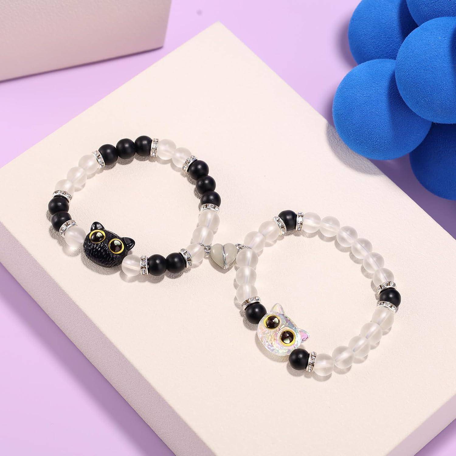 Zwei perlenbesetzte Armbänder mit Katzenkopf-Anhängern, eines schwarz und eines schillernd, auf einer hellrosa Schachtel vor einem lavendelvioletten Hintergrund präsentiert.
