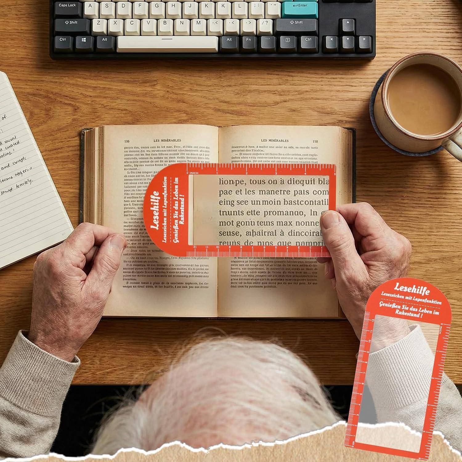 Hände halten ein rot durchsichtiges Lesehilfsmittel über ein aufgeschlagenes Buch auf einem Schreibtisch und vergrößern den Text zum leichteren Lesen; eine Tastatur und eine Kaffeetasse befinden sich in der Nähe.