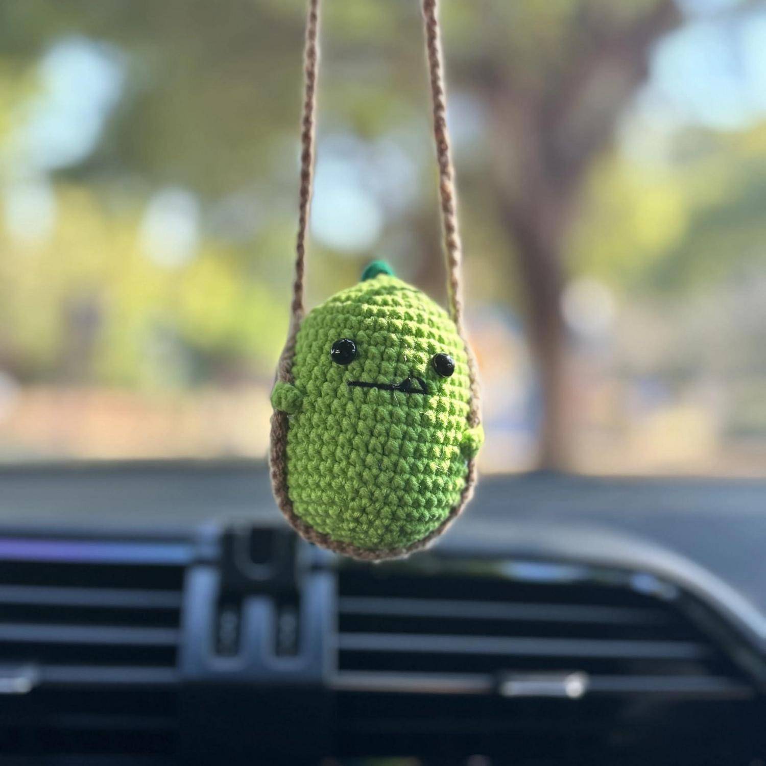 Kleiner grüner gehäkelter Kaktus, der an einem Rückspiegel eines Autos hängt, mit einem lächelnden Gesicht.