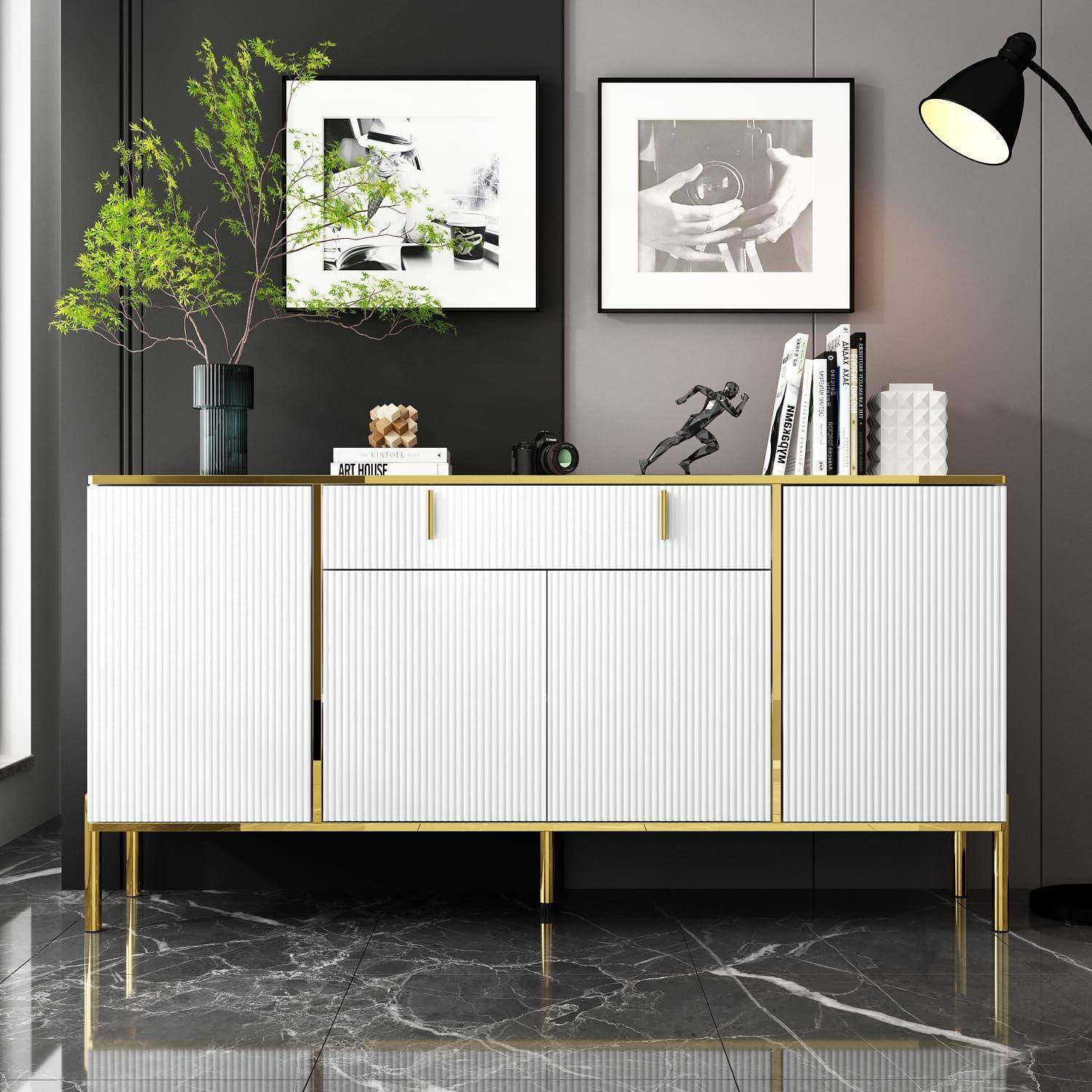 Modernes weißes Sideboard mit goldenen Beinen vor einer dunklen Wand, gekrönt mit einer Pflanze und Dekoration, darunter zwei gerahmte schwarz-weiße Bilder.