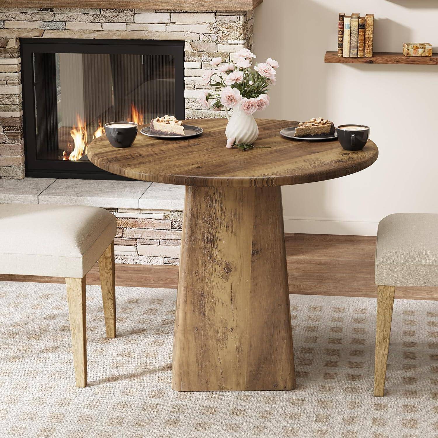 Rundes Holz-Esstisch-Set mit Tellern, Tassen und Blumen in einem gemütlichen Zimmer neben einem brennenden Kamin, flankiert von zwei gepolsterten Stühlen.
