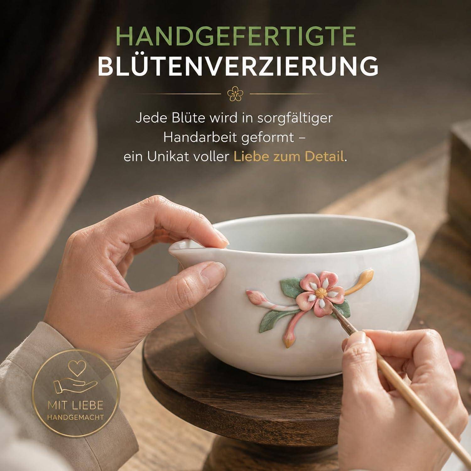 Hände verzieren eine Keramikschale mit einer kleinen Blütenapplikation; der deutsche Text lautet „HANDGEFERTIGTE BLÜTENVERZIERUNG" und besagt, dass jede Blüte sorgfältig von Hand angefertigt wird.