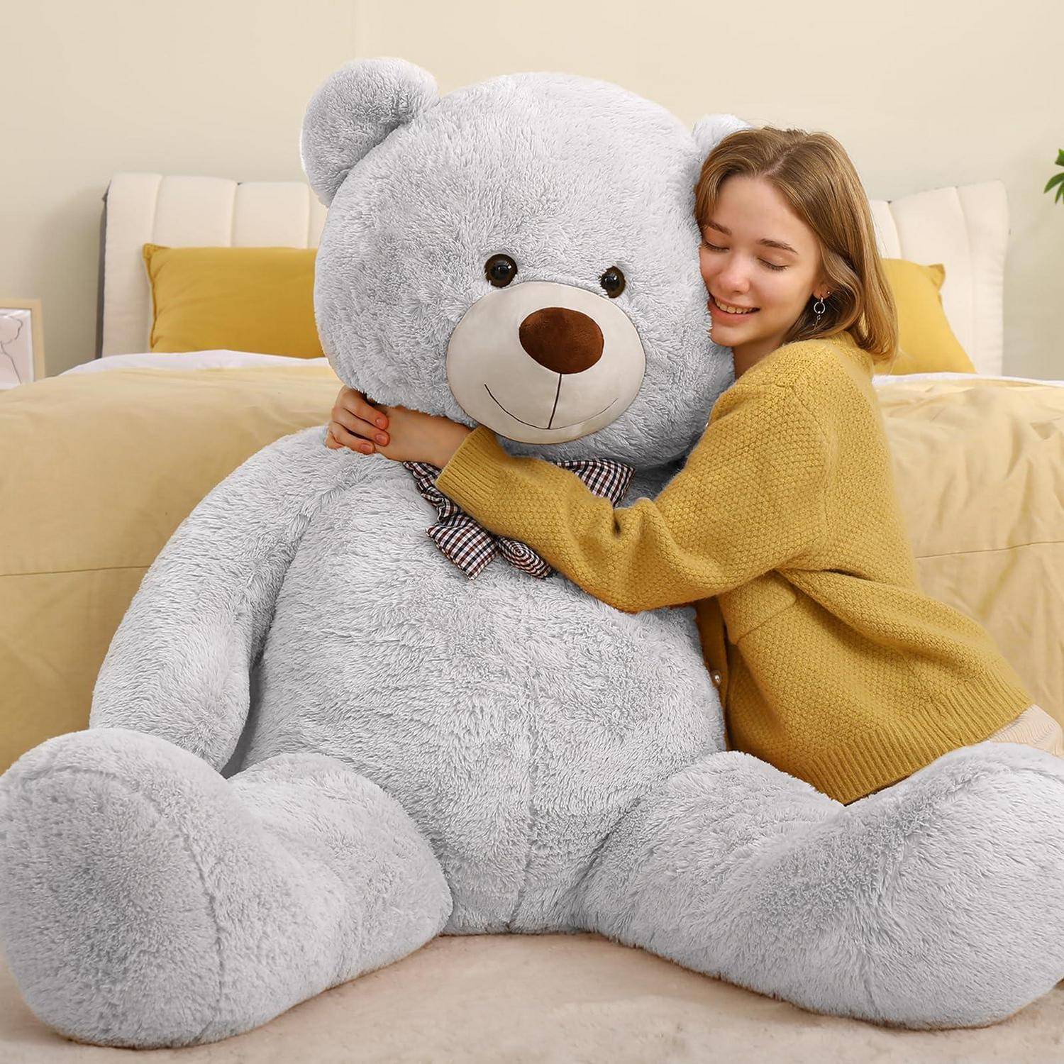 Junge Frau, die einen übergroßen grauen Teddybär auf einem Bett mit gelber Bettwäsche umarmt, lächelnd mit geschlossenen Augen.