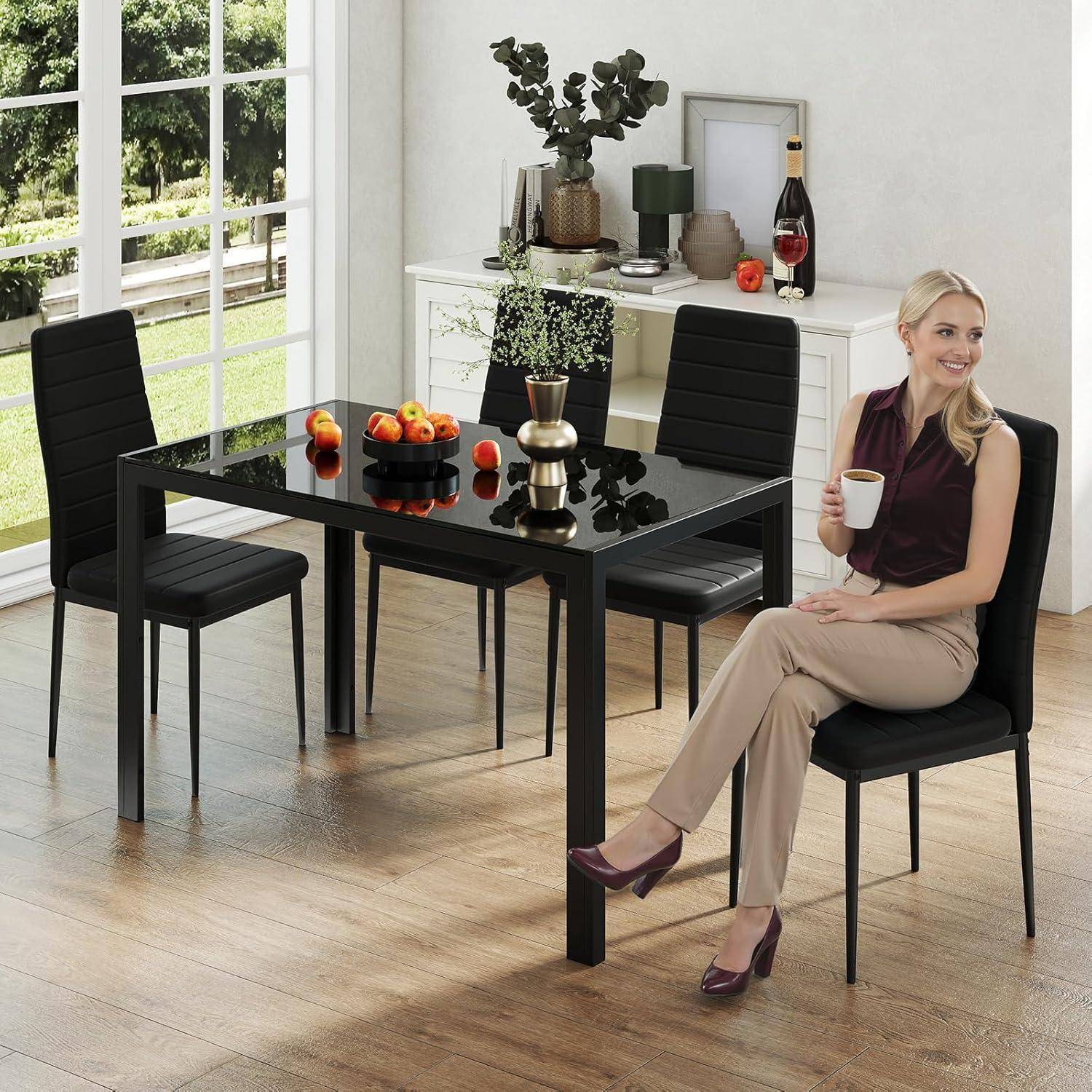 Blonde Frau sitzt an einem schwarzen Esstisch und hält eine Tasse in einem hellen Raum mit vier schwarzen Stühlen, Obstgesteck und einem Sideboard.