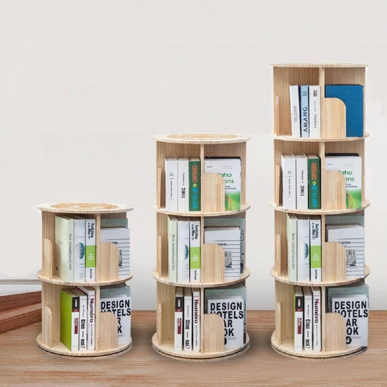 Drei kleine zylindrische Holzbücherregale mit zunehmender Höhe, jedes gefüllt mit Büchern, gestapelt auf einem Holzboden vor einer schlichten Wand.