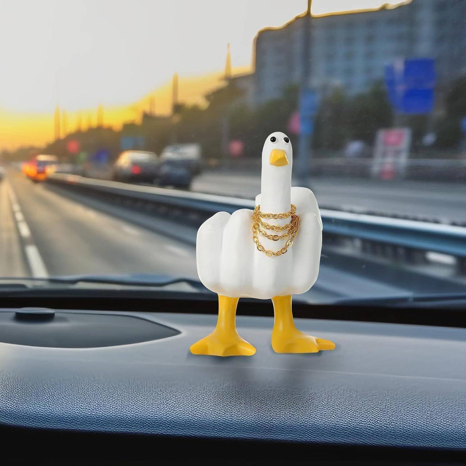 Ein kleine weiße Entenstatuette mit goldenen Ketten steht auf einem Armaturenbrett eines Autos, mit einer unscharfen Straße und Gebäuden einer Sonnenuntergangsstadt außerhalb der Windschutzscheibe.