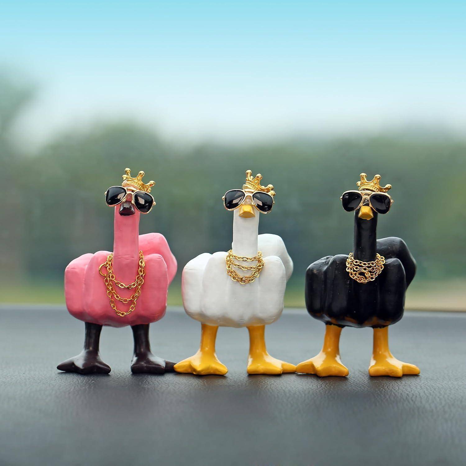 Drei kleine Vogelfiguren mit Kronen und Sonnenbrillen sowie goldenen Ketten in den Farben Rosa, Weiß und Schwarz, die nebeneinander im Freien stehen.