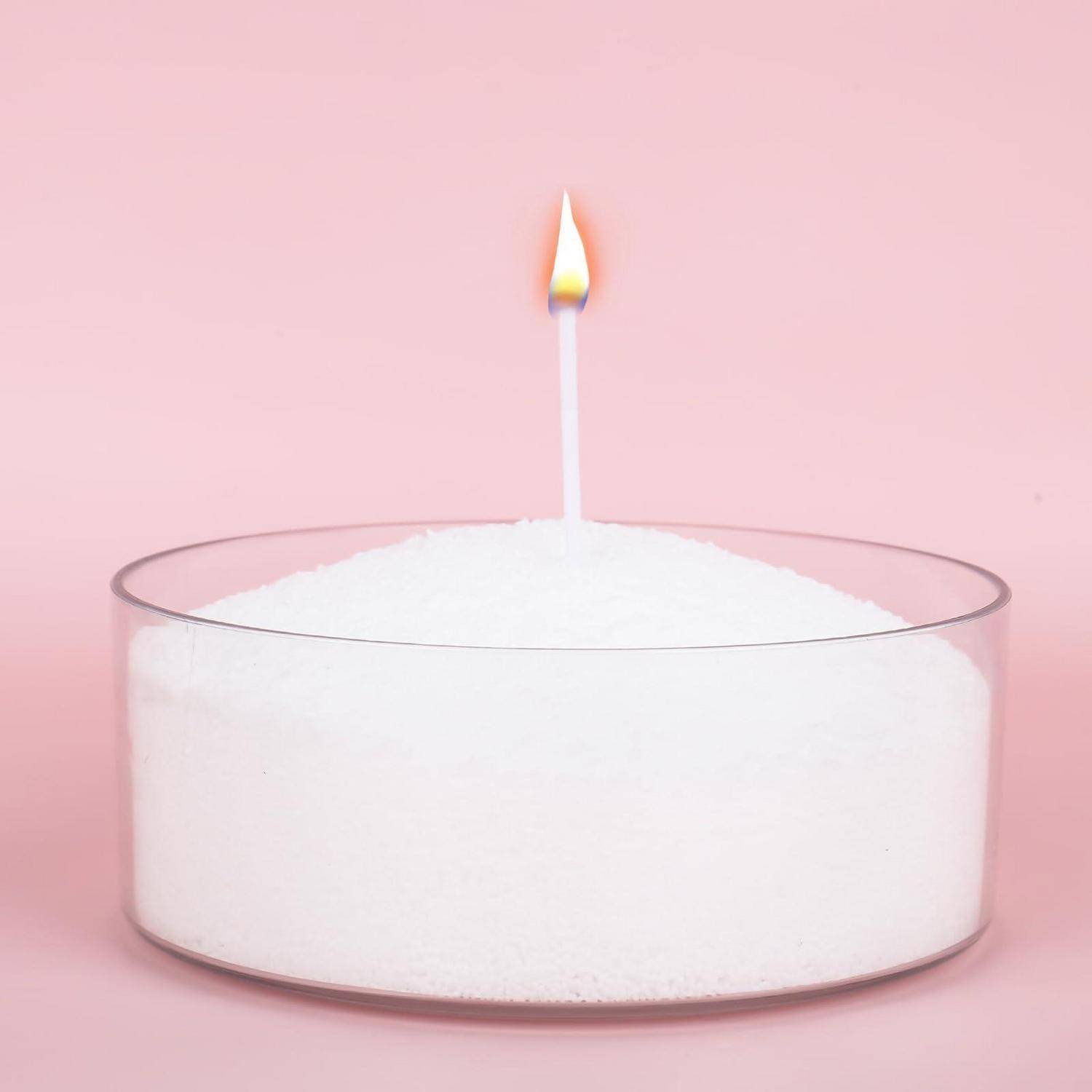 Eine einzelne brennende Kerze steht aufrecht in einem klaren Glasgefäß, das mit weißen Körnern gefüllt ist, vor einem rosa Hintergrund.