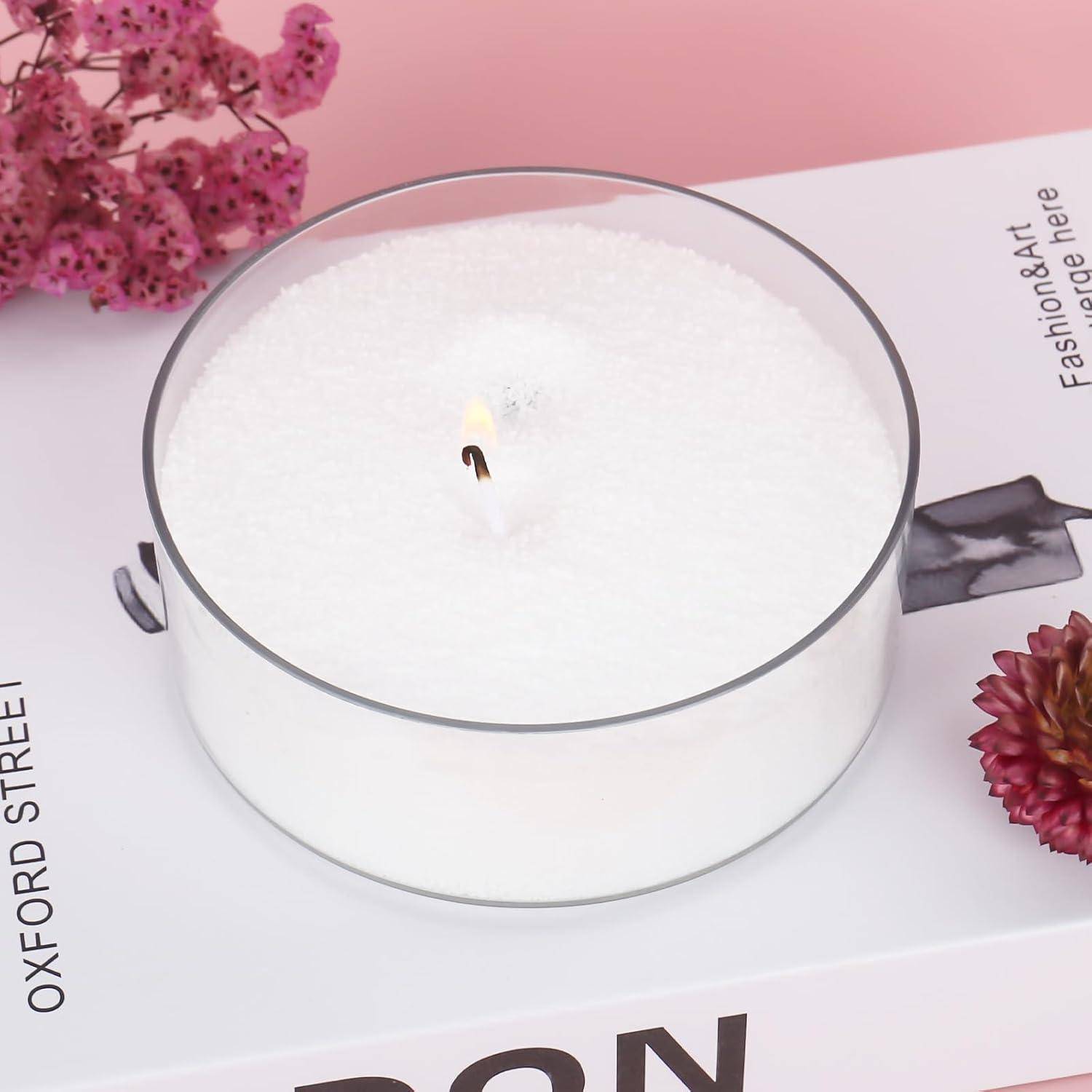 Eine weiße duftende Kerze in einem klaren Glaseimer mit einer kleine brennenden Docht, platziert auf einem rosa-weißen Buch neben getrockneten Blumen.