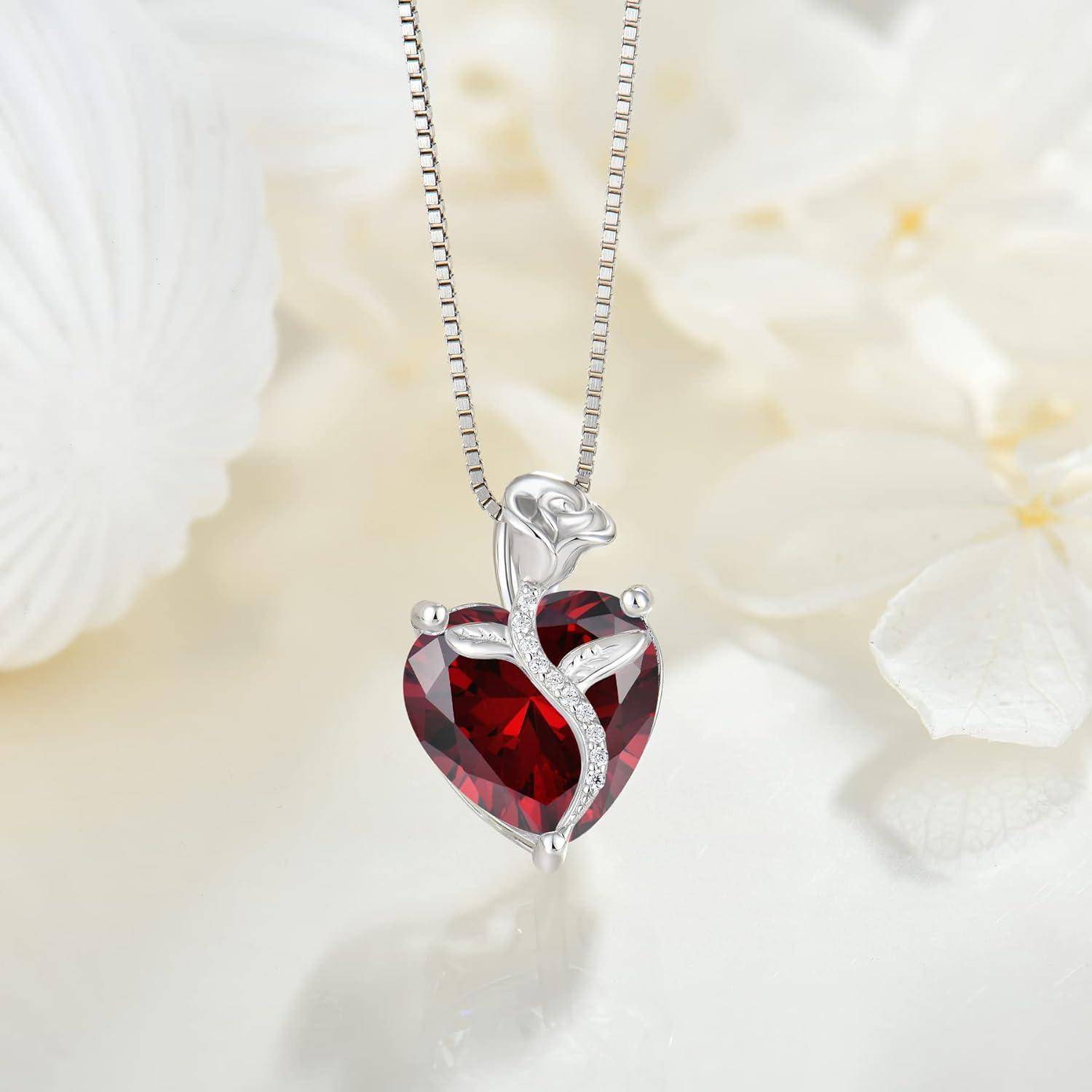 Silberner herzförmiger Anhängercollier mit einem roten Edelstein und kleinen klaren Steinen, hängend an einer Kette vor einem weichen weißen Blumenhintergrund.