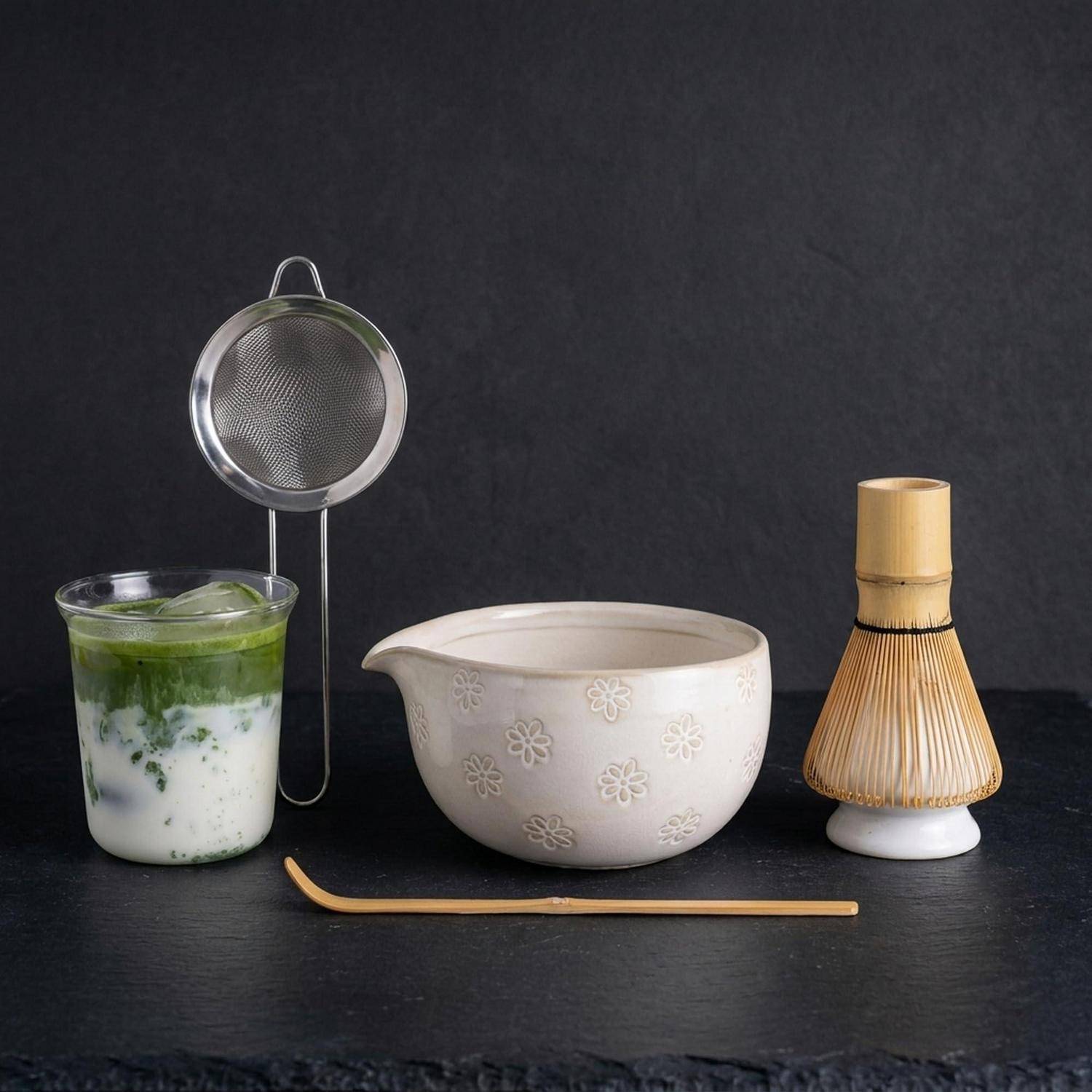 Matcha-Tee-Set mit einem Glas eines geschichteten grün-weißen Getränks, Schneebesen-Sieb, Keramikschale, Bambusbesen und Löffel auf dunklem Hintergrund.