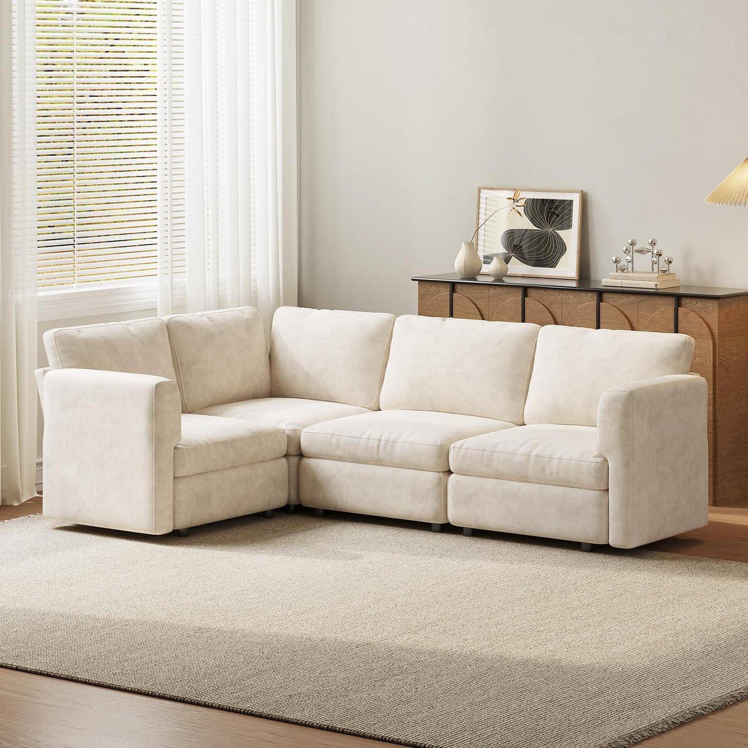 Cremefarbenes L-förmiges Ecksofa in einem hellen, modernen Wohnzimmer mit einem Teppich, Fensterjalousien und einem Holzkommode dahinter.
