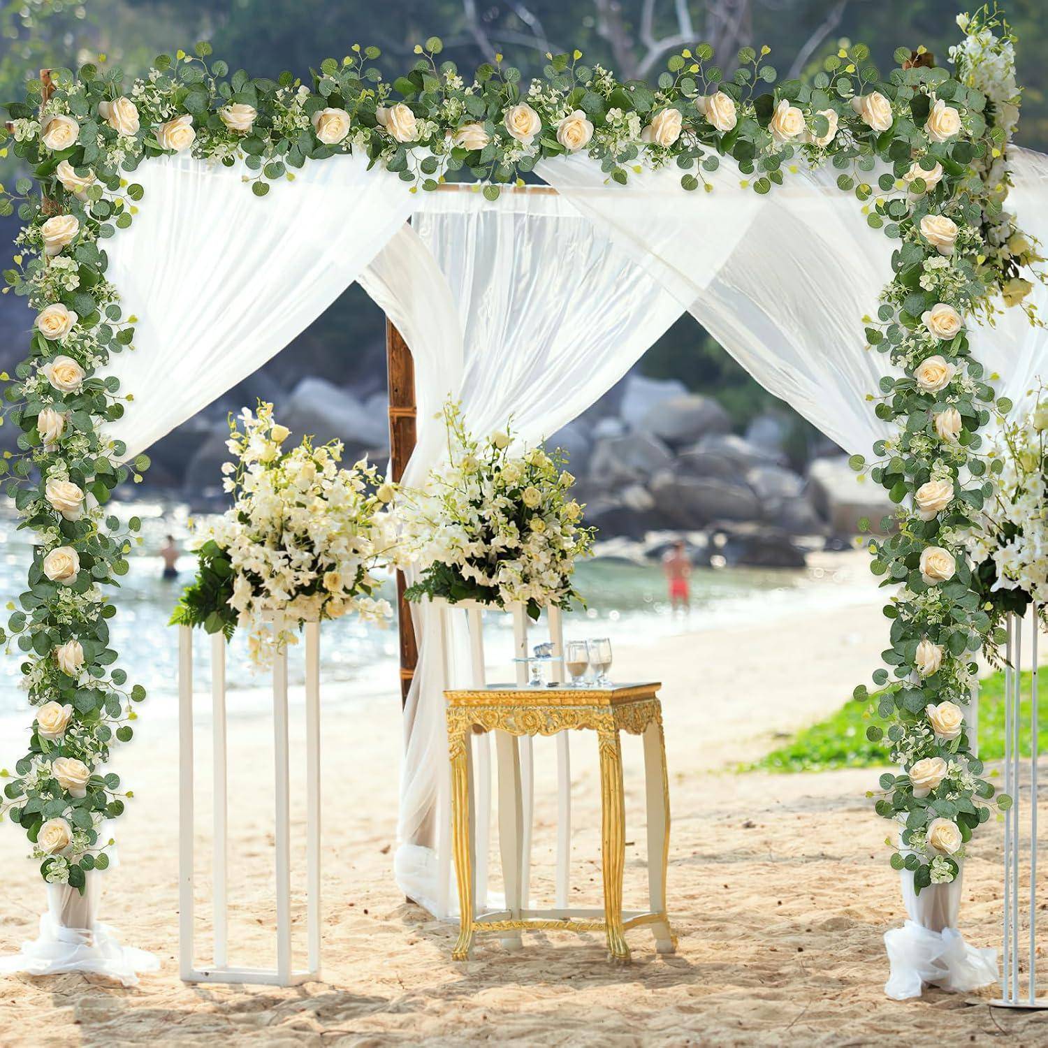 Blumenschmückter Traubogen für eine Strandtrauung mit weißen Drapierungen, Blumenständern und einem kleinen Goldtisch im Sand am Meer.