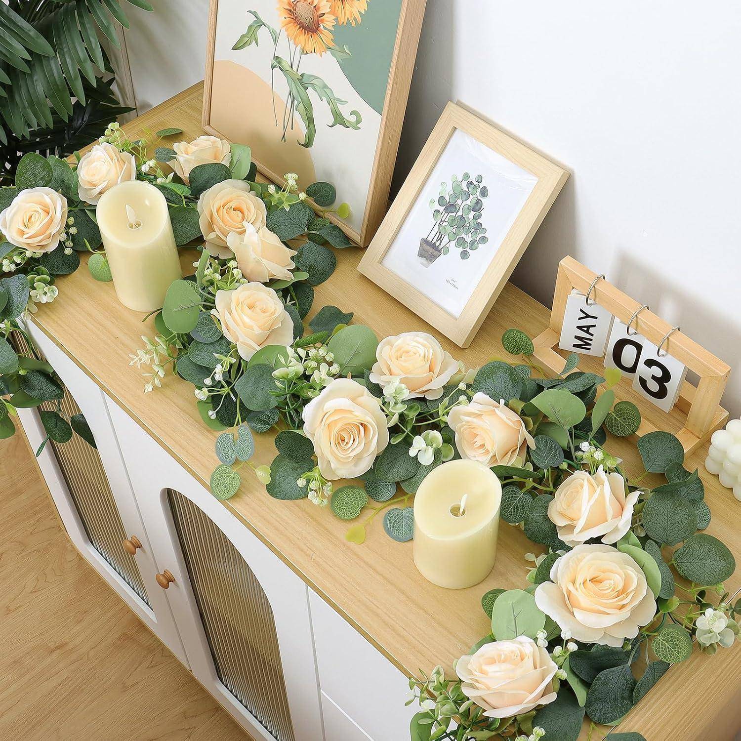 Dekorierter Holzschrank mit cremefarbenen Rosen, Grünflächengirlande, Kerzen, gerahmter Blumenkunst und einem Schreibtischkalender vom 03. Mai.