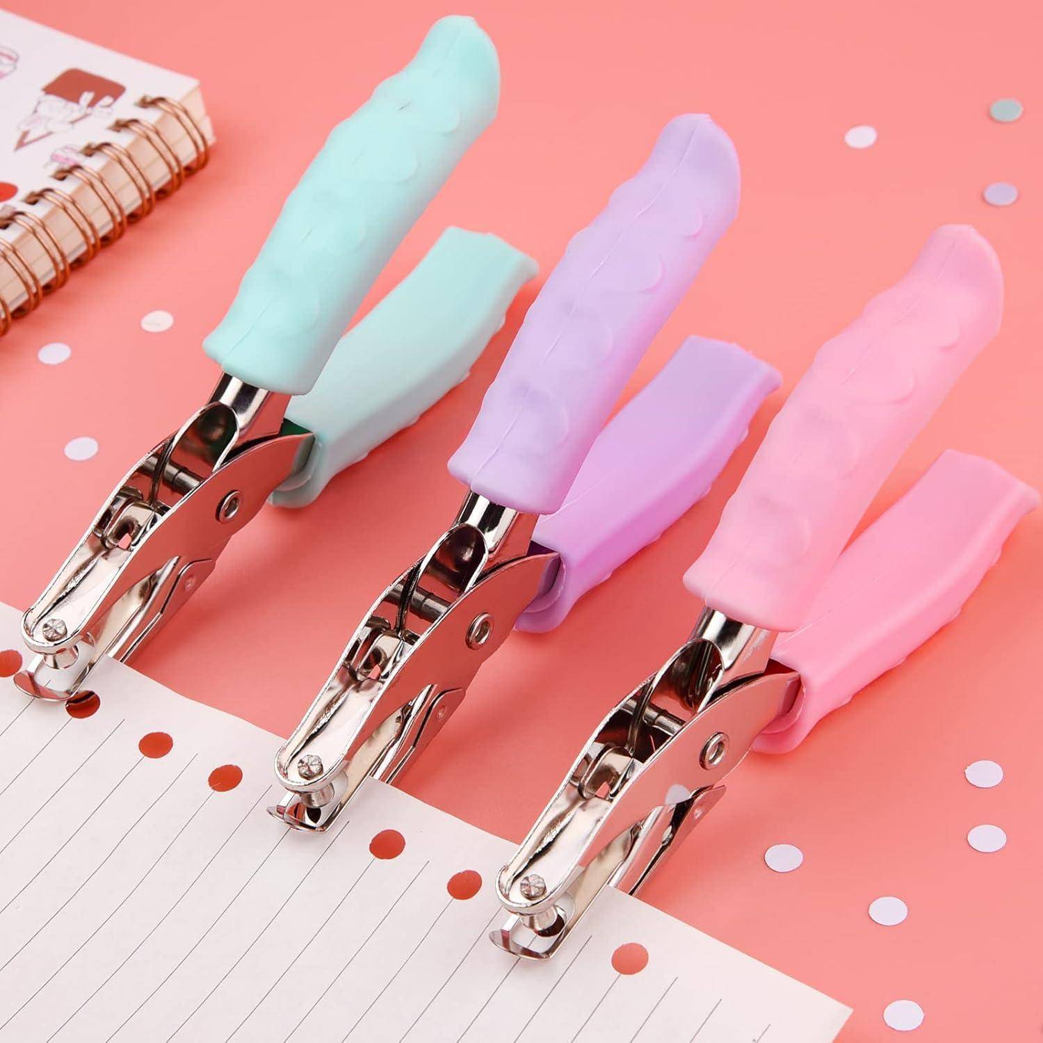 Drei Pastelllocheisen mit Gummigriff auf einem rosa Schreibtisch neben einem linierten Notizbuch mit Konfetti-Punkten.