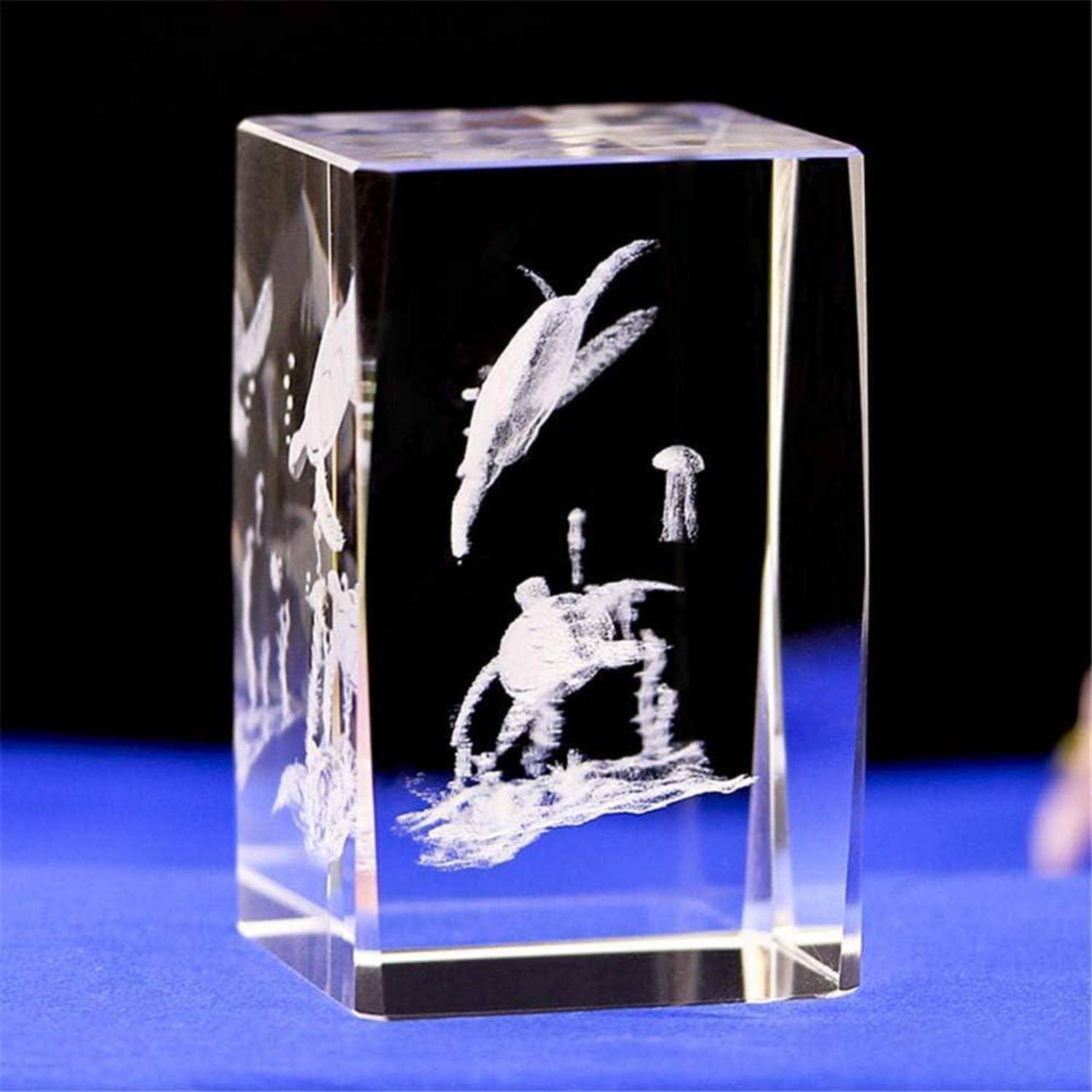 Ein klarer Glasblock mit eingravierten Meerestieren, darunter ein Delphin, Fische und eine Krabbe, ausgestellt auf einer blauen Oberfläche vor schwarzem Hintergrund.