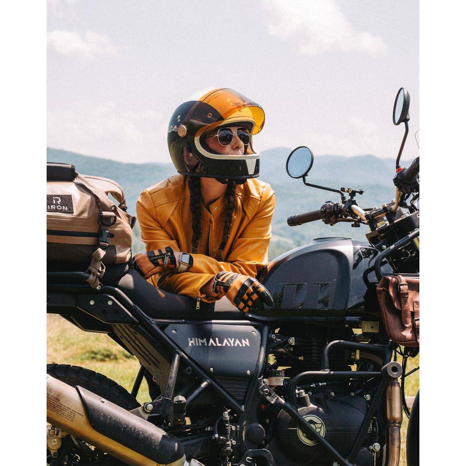 Ein Motorradfahrer mit Helm in einer gelben Jacke sitzt auf einem Himalayan-Motorrad und lehnt sich auf den Lenker, mit Bergen im Hintergrund.
