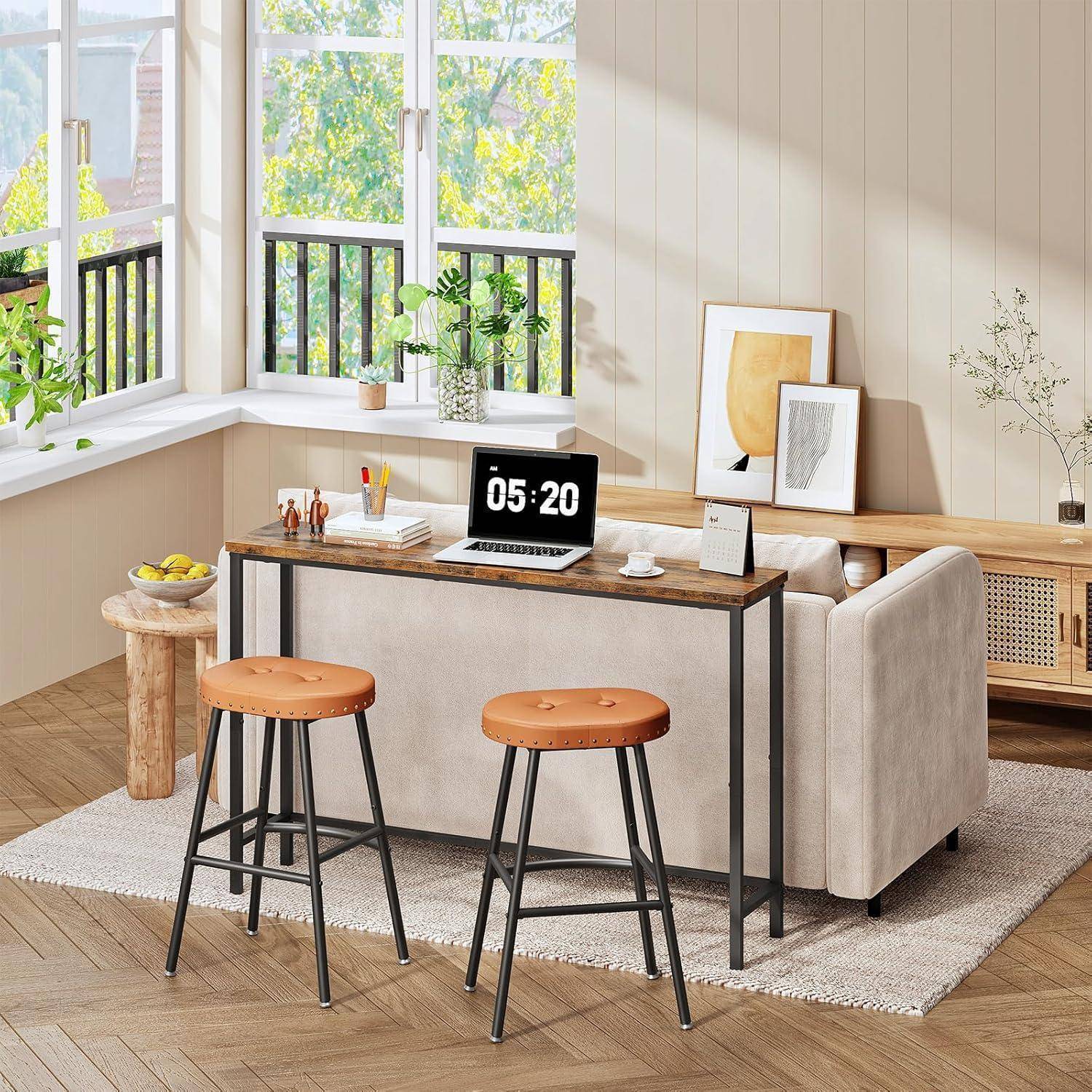Helles modernes Wohnzimmer mit zwei Hockern an einem schmalen Schreibtisch, aufgeklapptem Laptop mit Anzeige 05:20, Sofa, Pflanzen und gerahmter Kunst neben großen Fenstern.