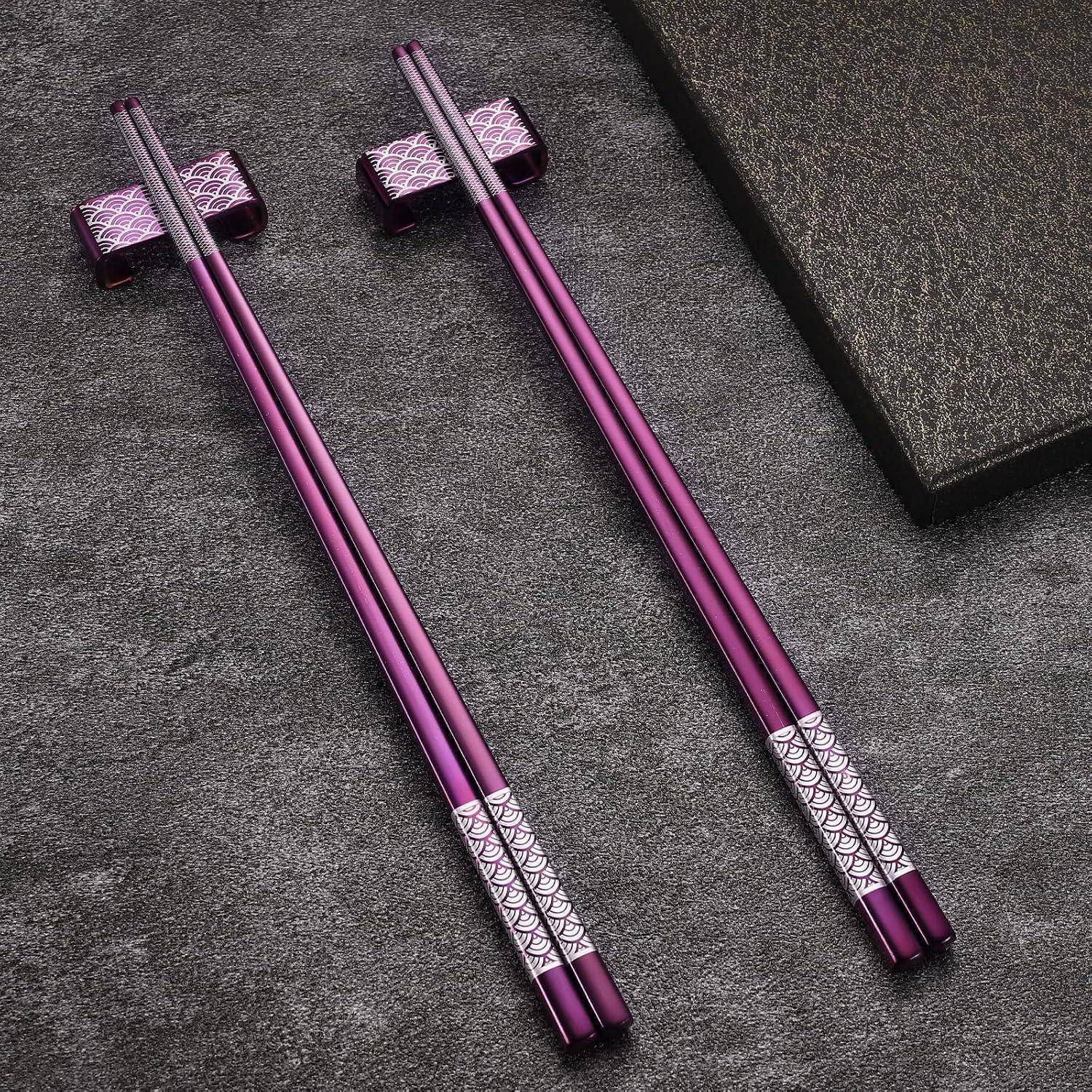 Zwei Paar violette Metallstäbchen mit gemusterten Griffen und passenden Haltern auf einer dunkel strukturierten Oberfläche.