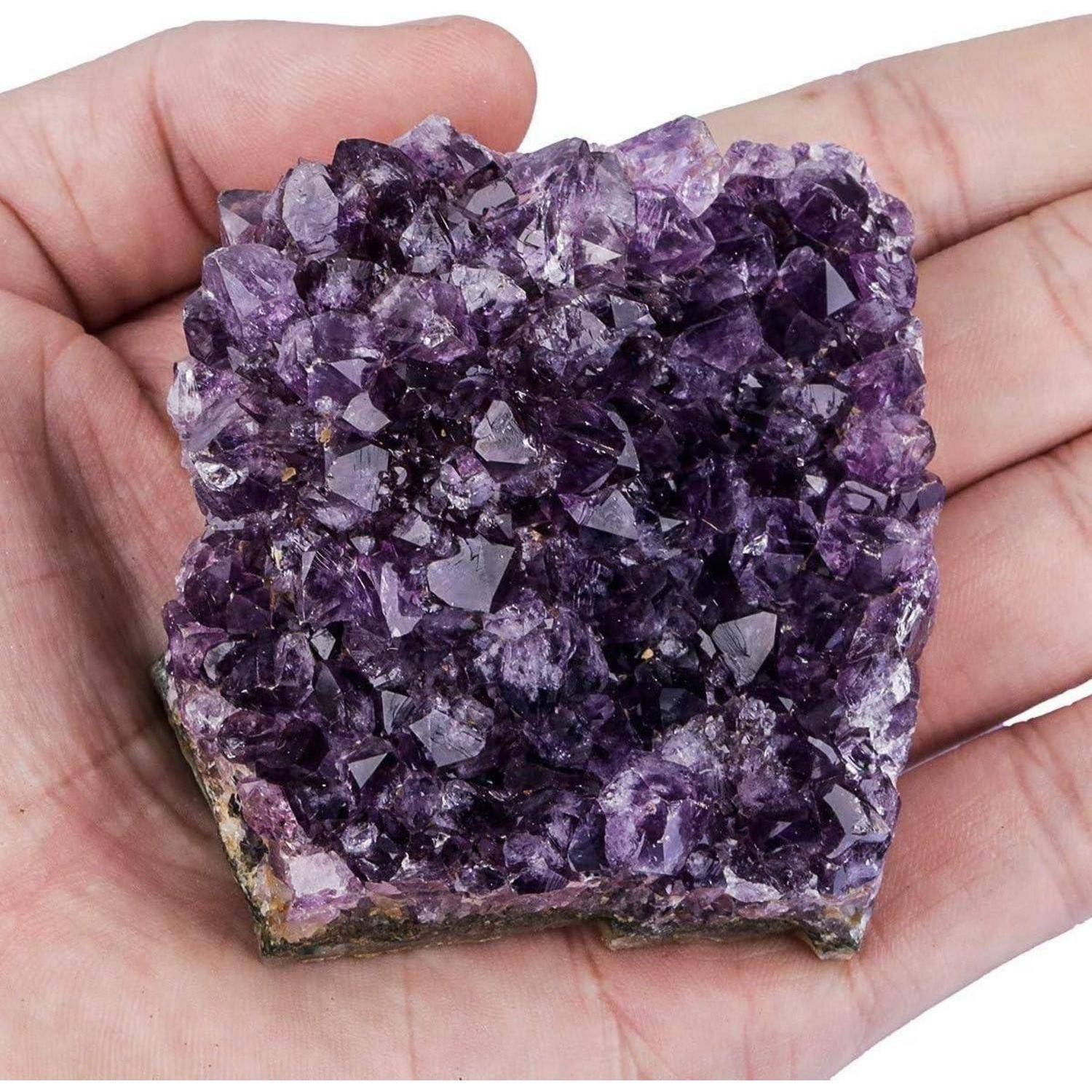 Eine Hand hält eine violette Amethyst-Kristalldruse mit spitzen funkelnden Kristallen auf einem kleinen Felssockel.