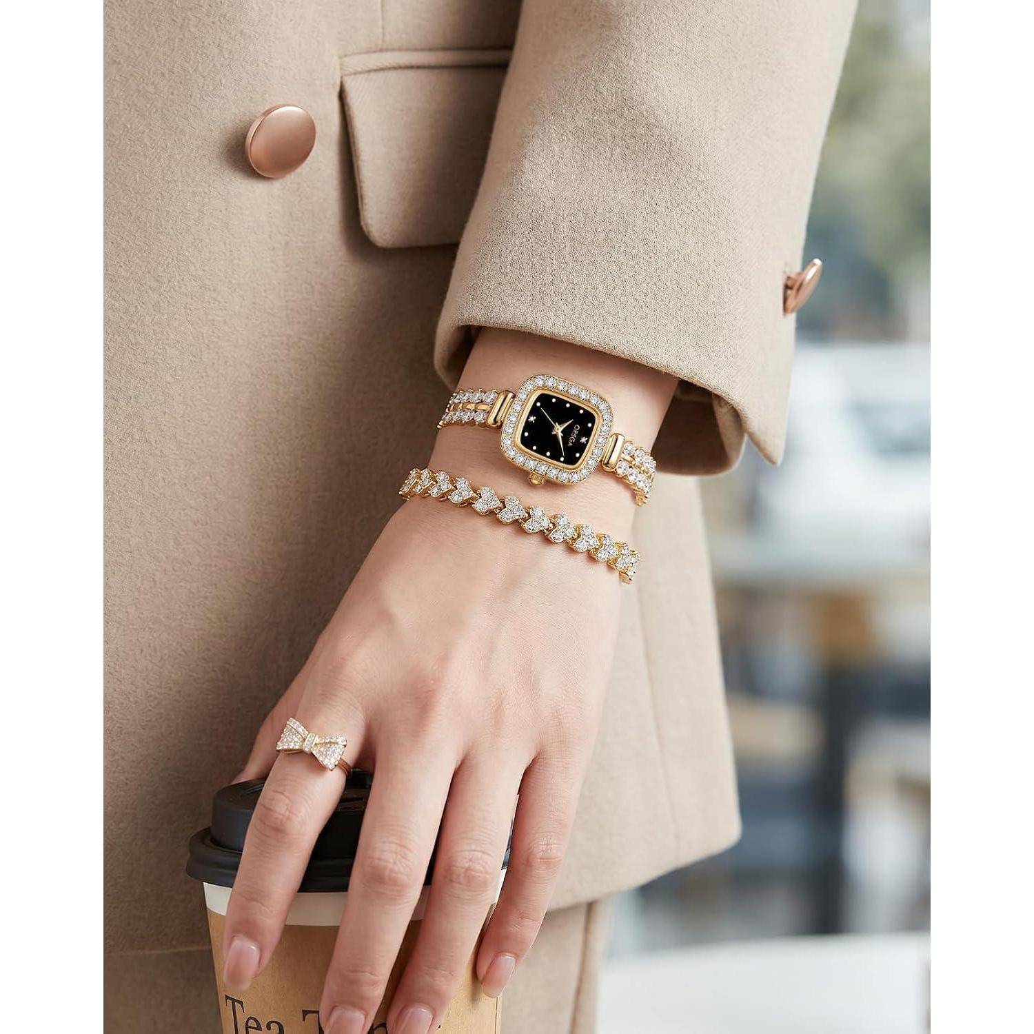 Nahaufnahme einer Hand, die eine Kaffeetasse hält, mit einer goldenen Uhr, Armbändern und einem Ring, während ein Ärmel eines beigefarbenen Mantels sichtbar ist.