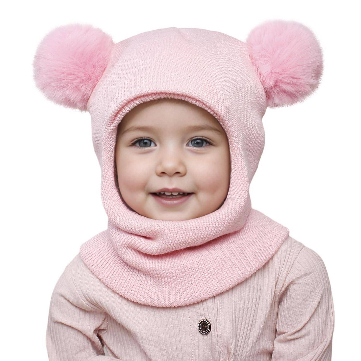 Lachendes Kleinkind in einer rosa Strickmütze und passendem Schal mit flauschigen Pompon-Ohren, vor weißem Hintergrund gezeigt.