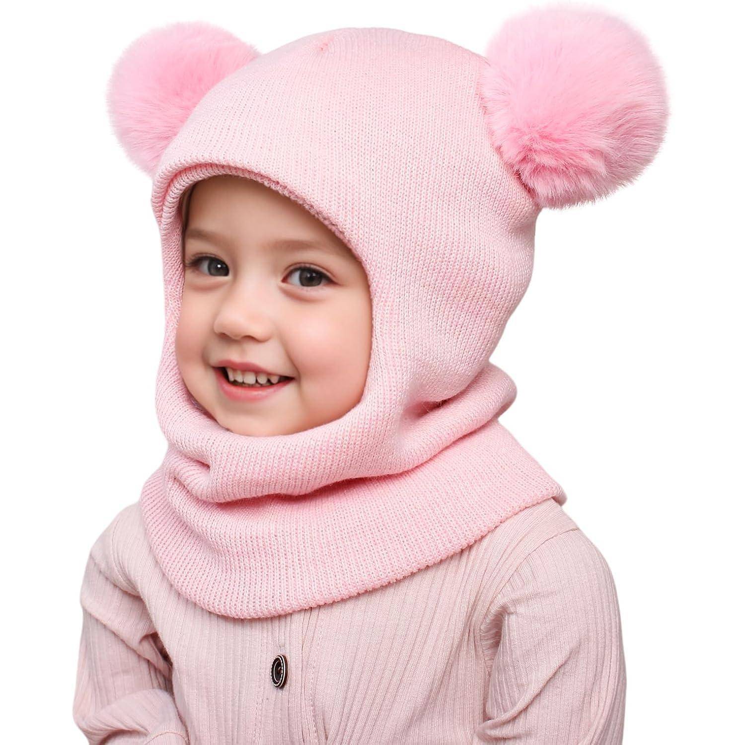 Lachendes junges Kind in einer rosa Strickmütze und passendem Schal mit flauschigen Pompons auf weißem Hintergrund.