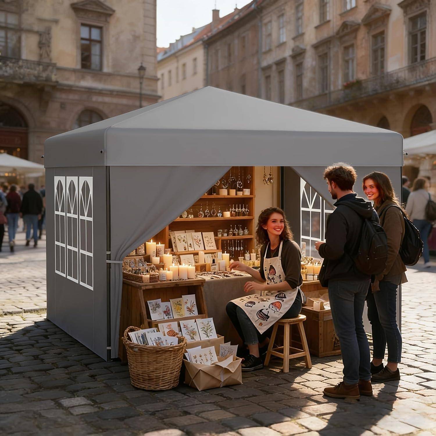 Eine Frau verkauft handgefertigte Karten und Kerzen von einem Marktstand aus, während zwei Kunden in einem sonnigen Kopfsteinplaster-Platz stöbern.