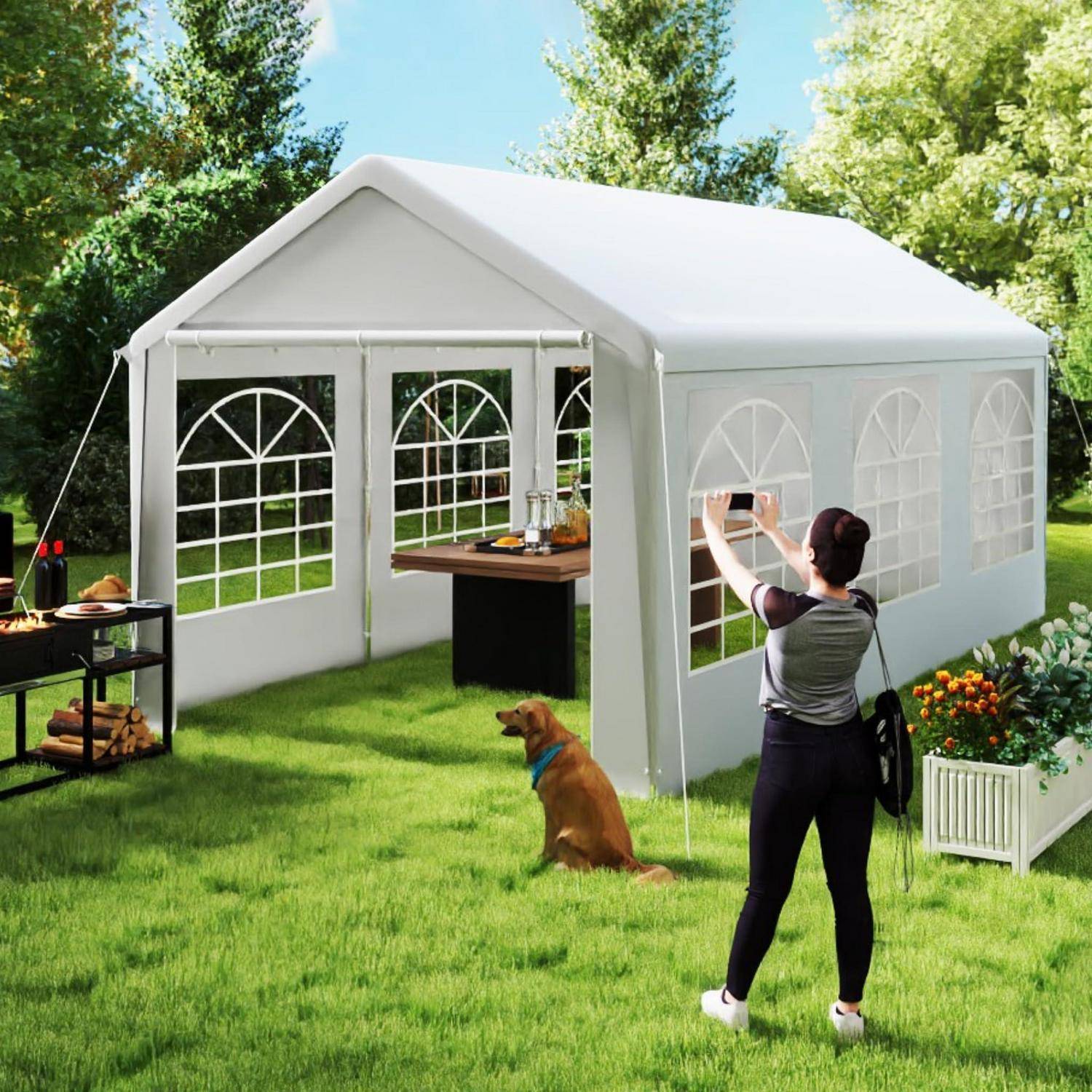 Eine Person baut ein weißes Outdoor-Pavillon-Zelt auf einer Wiese im Hinterhof auf, mit einem Hund in der Nähe und einem kleinen Tisch sowie einem Grill unter dem Zelt.