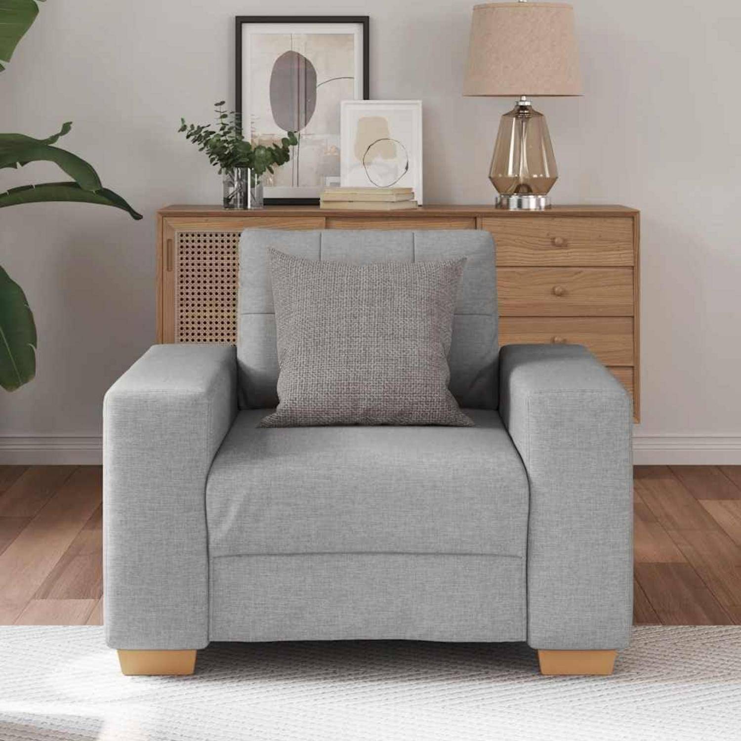 Hellgrauer gepolsterter Sessel mit passendem Kissen in einem modernen Wohnzimmer, vor einer Holzkommode mit Lampe und gerahmtem Kunstwerk.