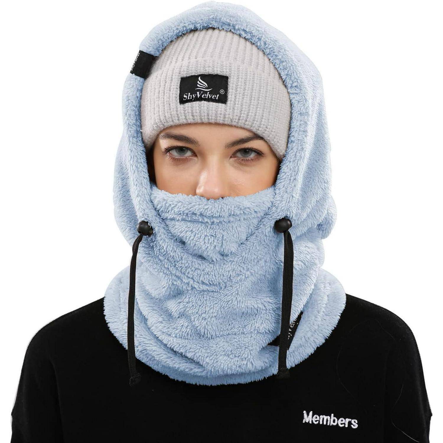 Person trägt eine hellblaue Fleece-Sturmhaube und eine graue Mütze, die Kopf und Hals bedecken, wobei nur die Augen sichtbar sind, vor einem weißen Hintergrund.