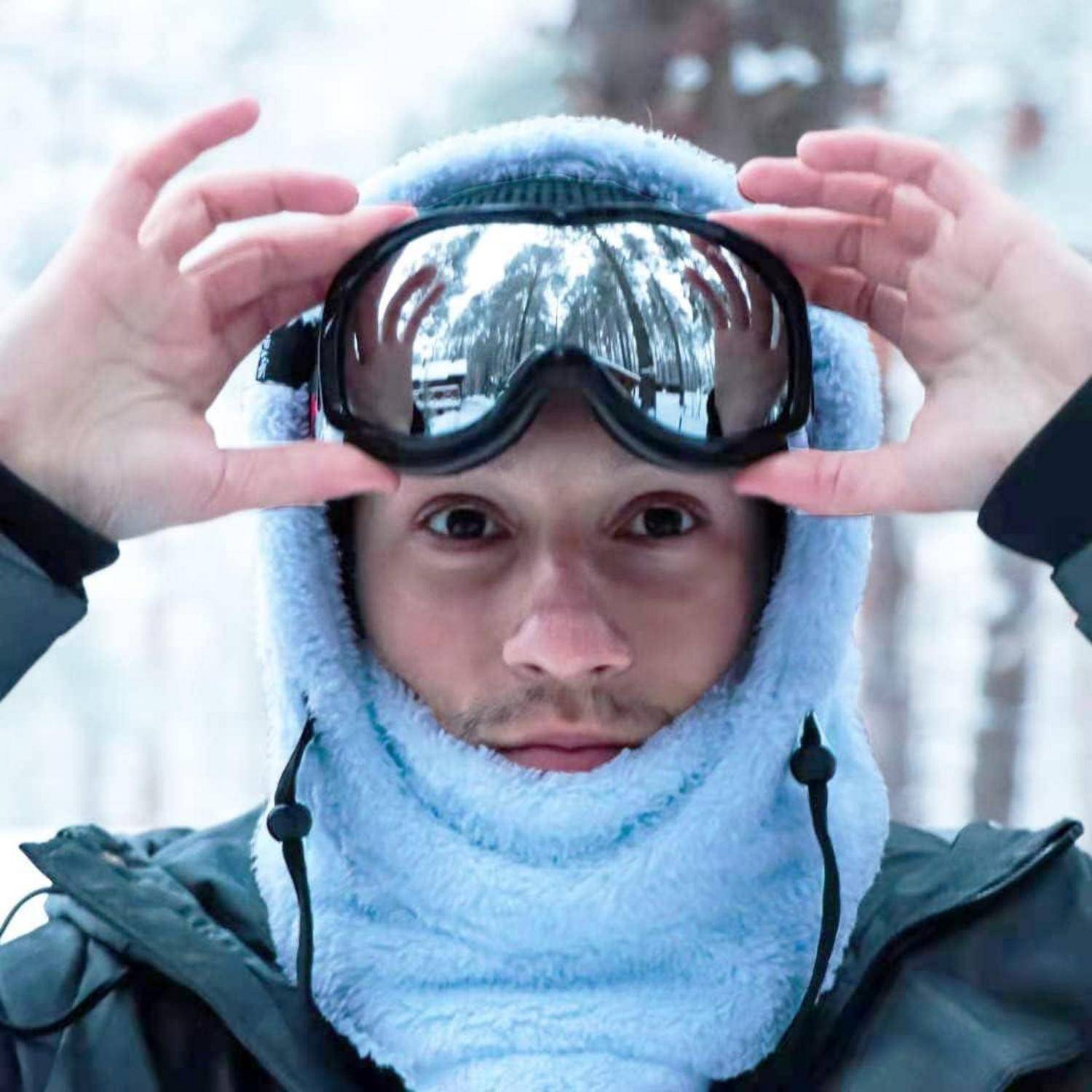 Nahaufnahme einer Person in Winterausrüstung, die Skibrillen und eine Gesichtsbedeckung in einem verschneiten Wald anpasst.