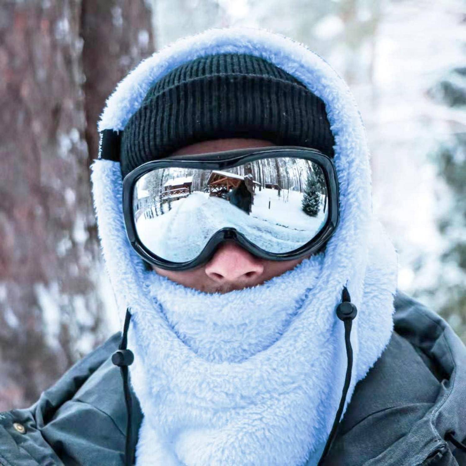 Nahaufnahme einer Person, die in Winterkleidung gehüllt ist, Skibrillen trägt, eine Kapuze und eine Gesichtsbedeckung in einer verschneiten Außenumgebung trägt.
