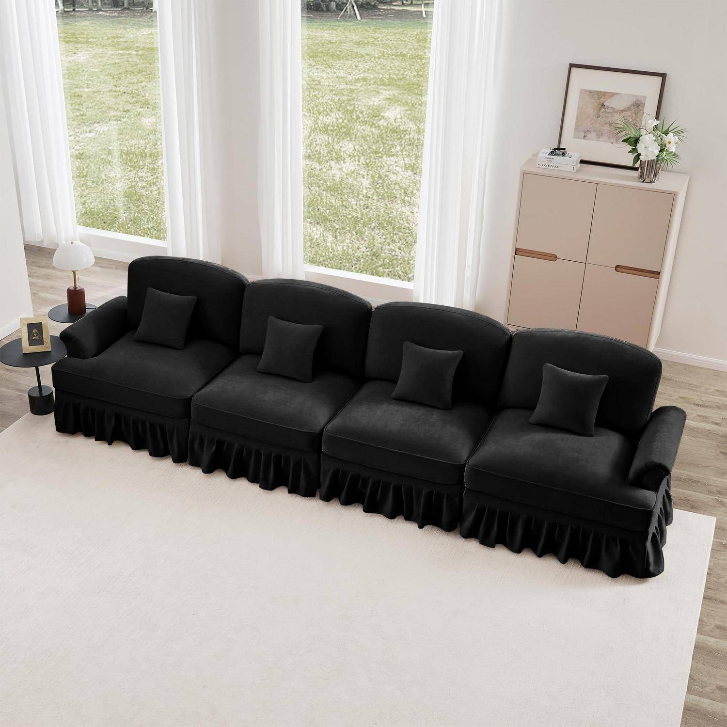 Schwarzes viersitziges Sofa mit kleinen Kissen in einem hellen Wohnzimmer, platziert vor hohen Fenstern und einem beigen Schrank mit Dekoration.