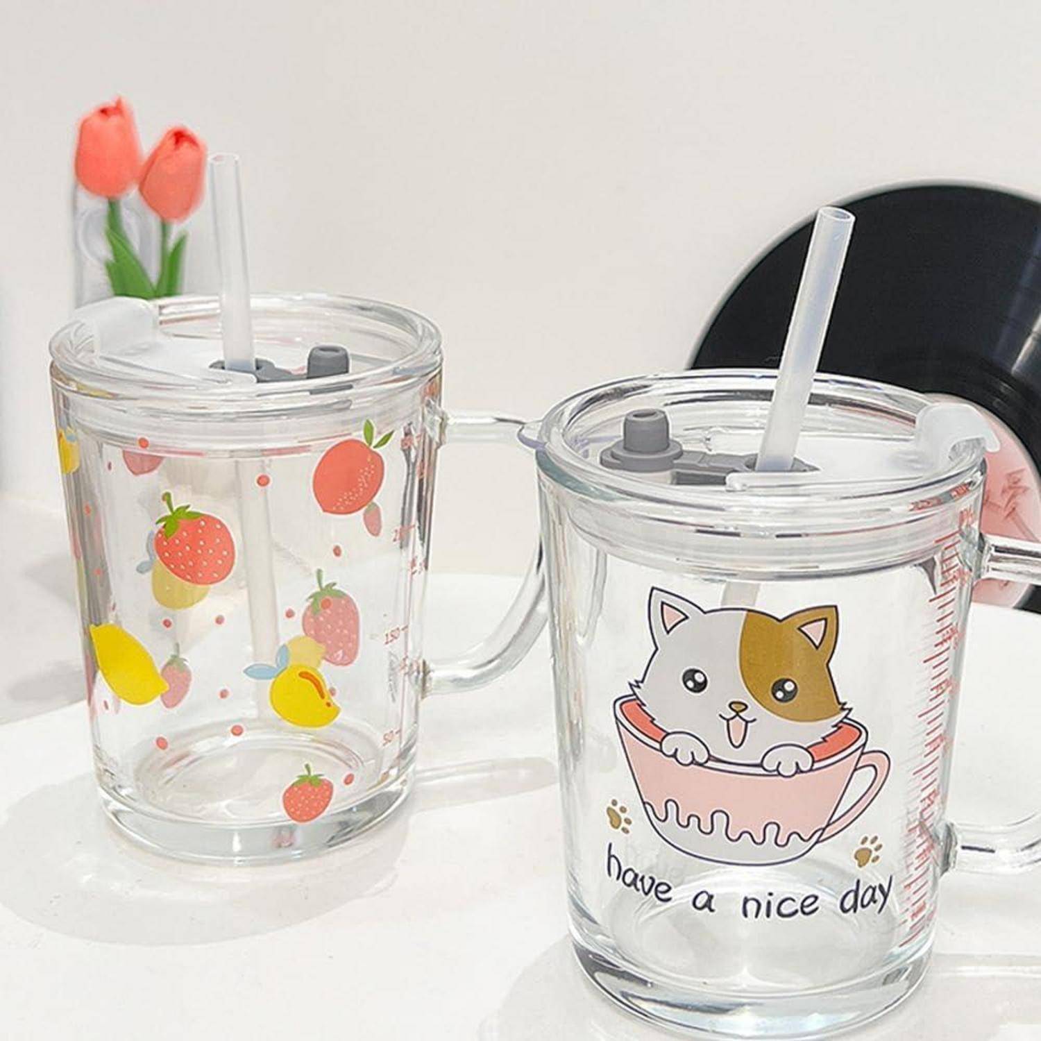Zwei durchsichtige Trinkbecher mit Griffen, Deckeln und Strohhalmen auf einer weißen Oberfläche, einer mit Erdbeeren- und Zitronenmustern, der andere mit einer Katze und dem Text „have a nice day