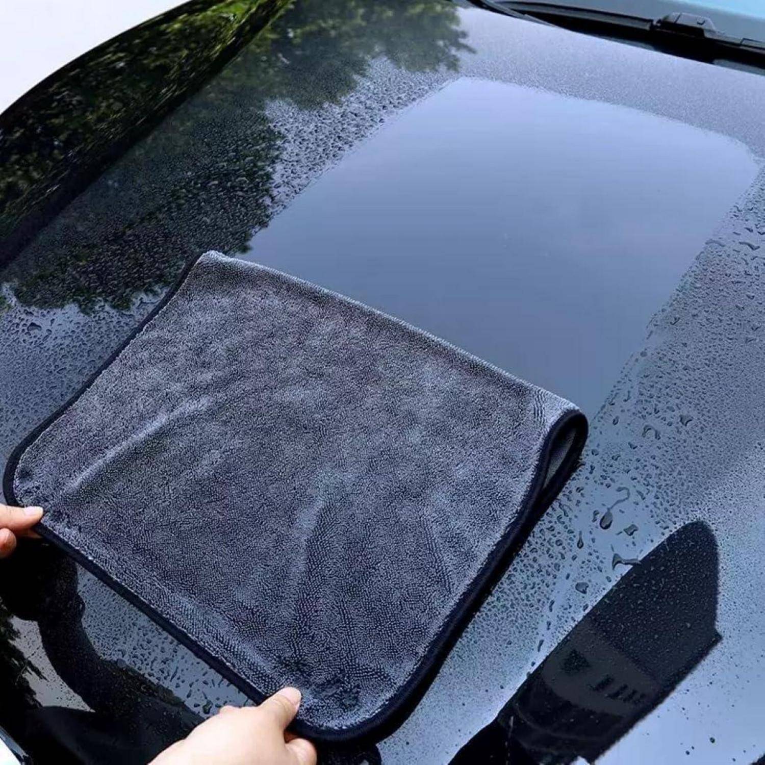 Eine Person hebt ein gefaltetes graues Mikrofaser-Handtuch von einer nassen Autowindschutzscheibe auf, was auf Trocknen oder Reinigung nach Regen hindeutet.