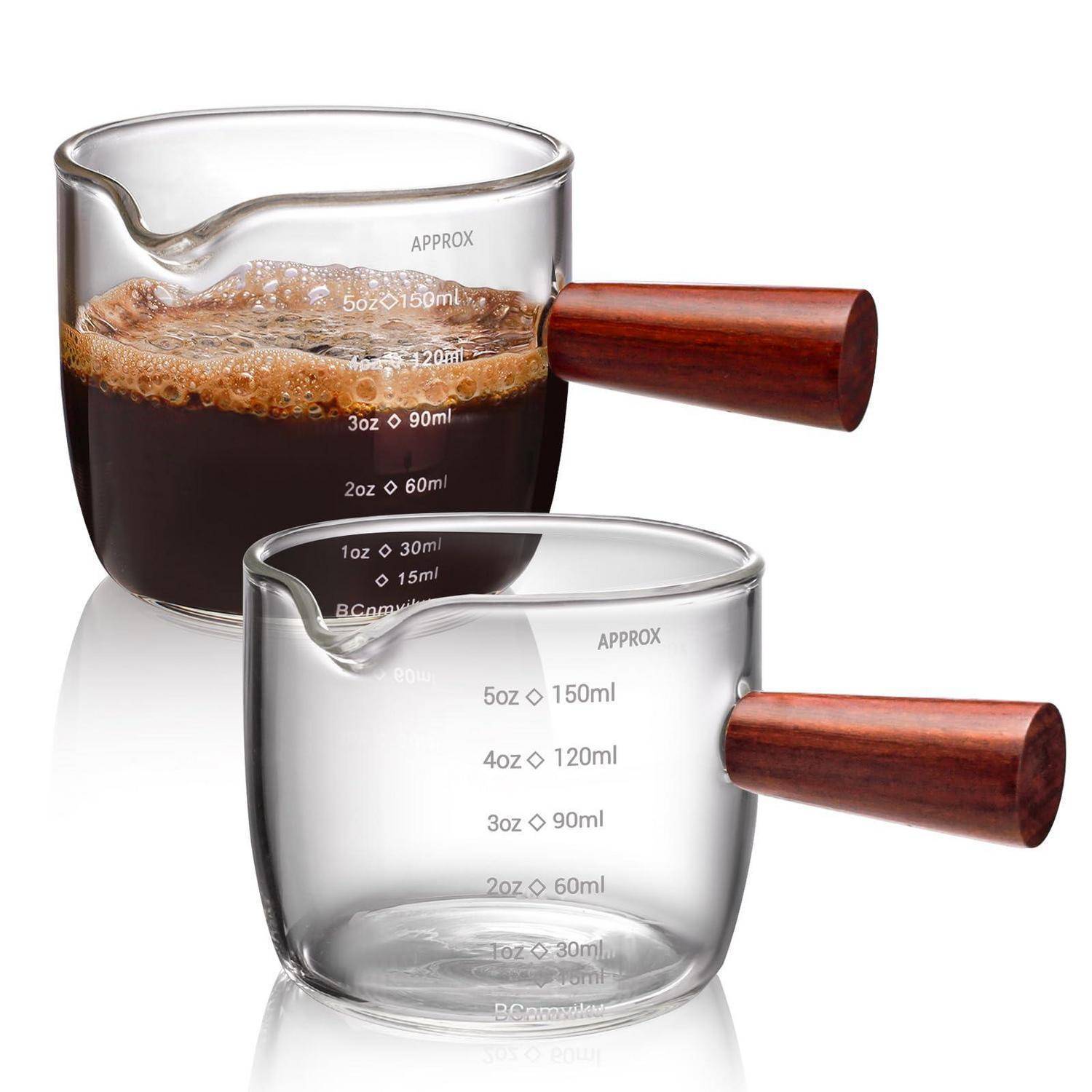 Zwei Glas-Kaffee-Messbecherbecher mit Holzgriffen, einer gefüllt mit dunklem Kaffee und Schaum, der andere leer, mit Unzen- und Milliliter-Markierungen.