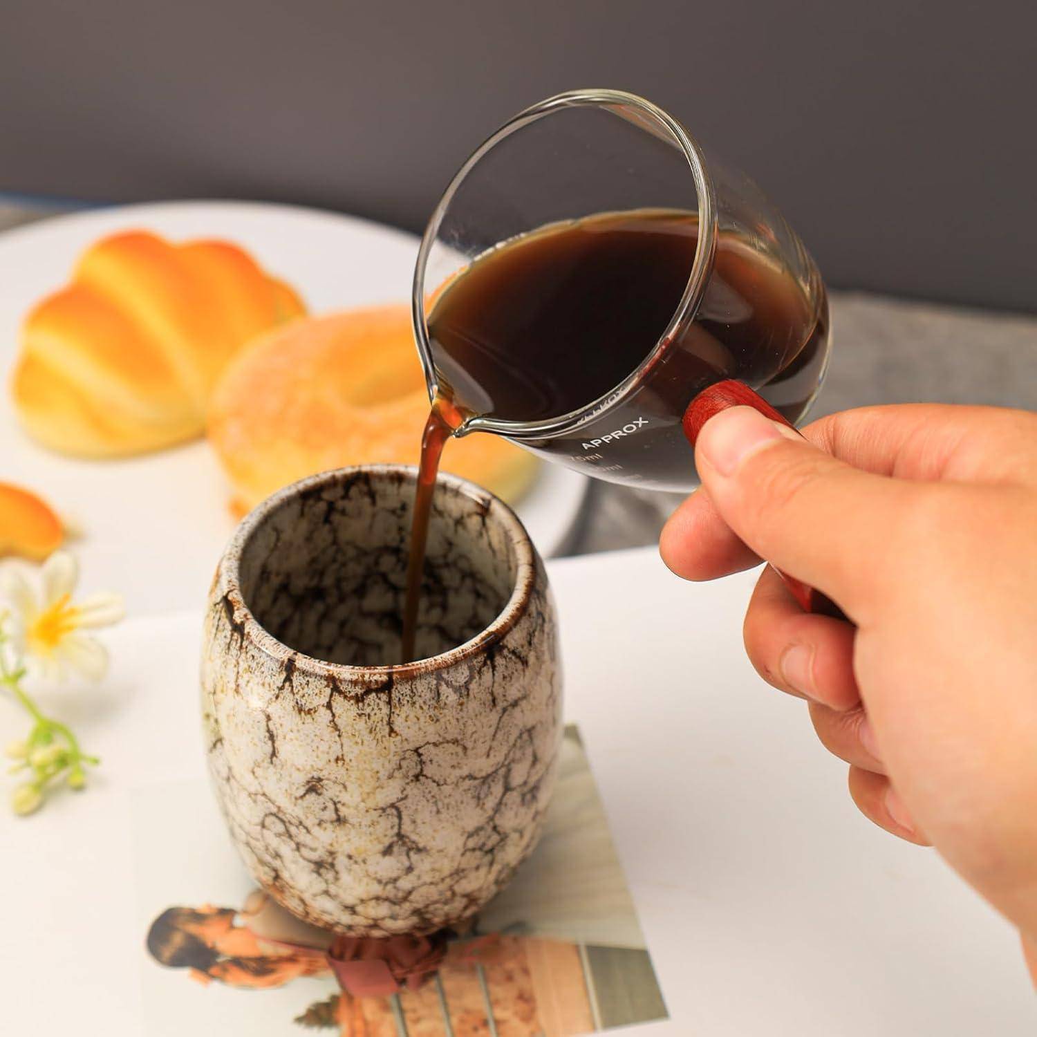 Eine Hand gießt dunklen Kaffee aus einer Glastasse in einen rustikalen Keramikbecher auf einem Tisch mit Gebäck und Blumendekoration.