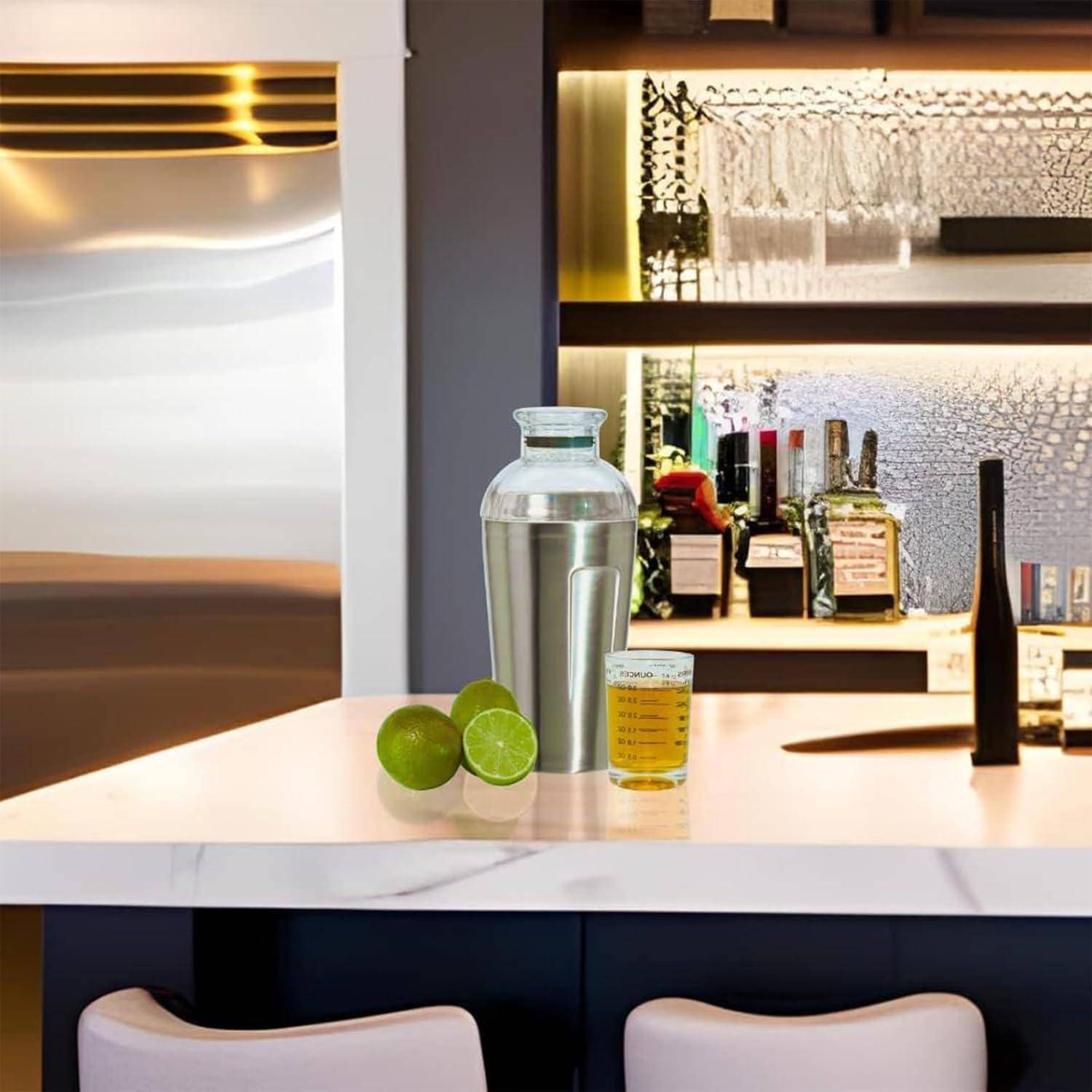 Stilvolle Theke mit einem Cocktail-Shaker, Limette und kleinem Trinkglas vor hinterbeleuchteten Regalen mit Flaschen und Gläsern.