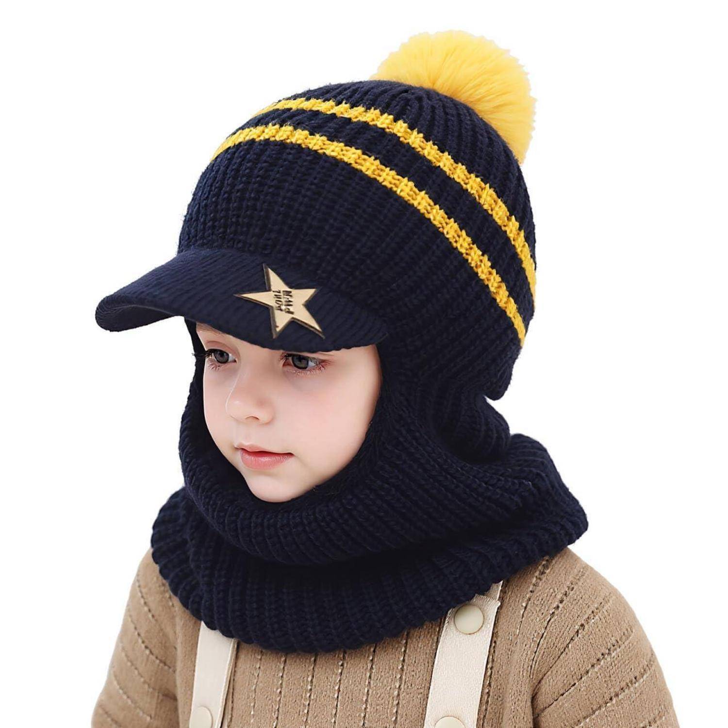 Kleines Kind mit einer dunkelblau gestrickten Wintermütze mit gelben Streifen, Bommel und angebrachtem Schirm, sowie passendem Schal-Kapuzen-Set und beigem Pullover.